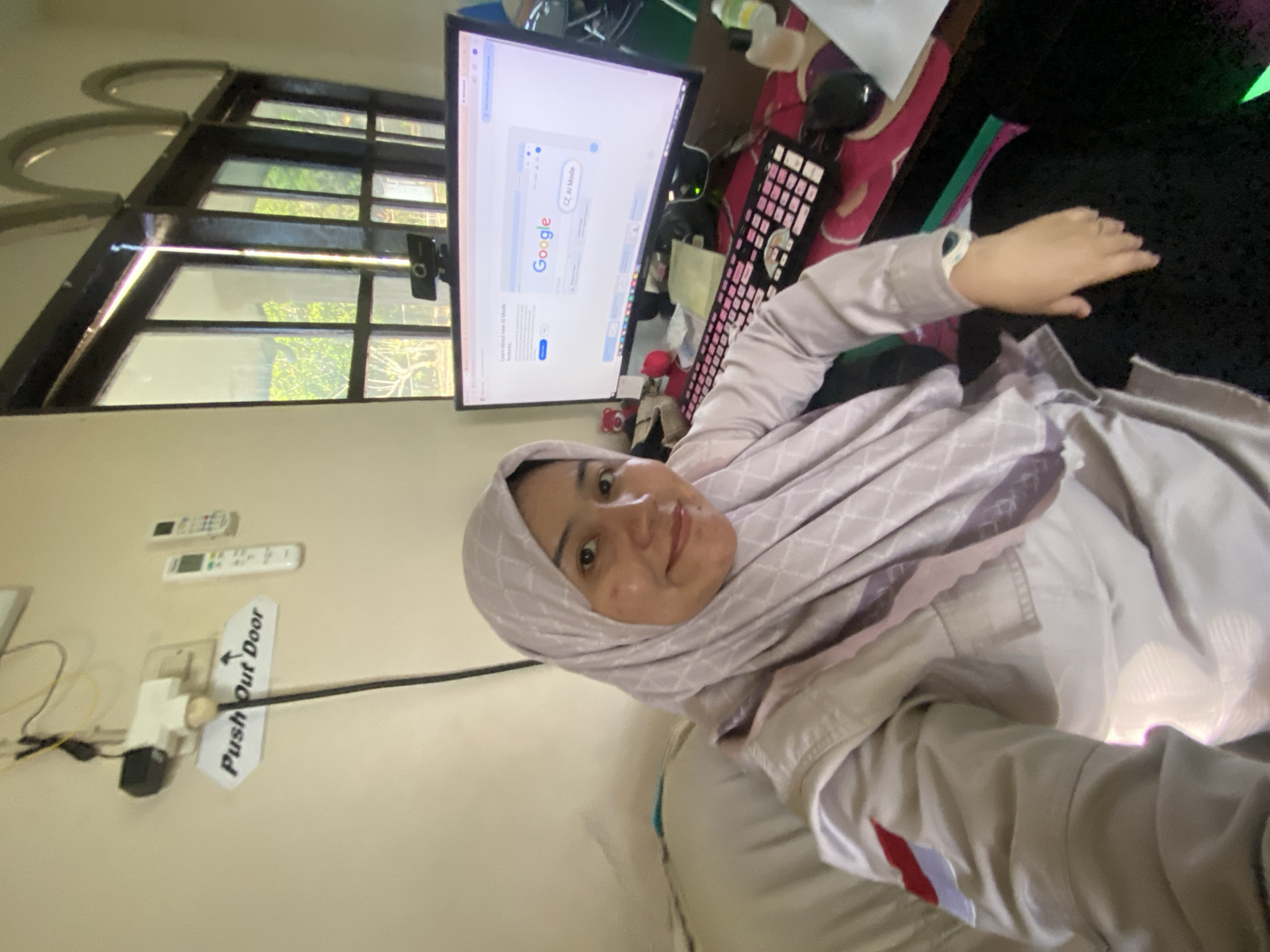 The image depicts a young woman wearing a hijab and a light-colored jacket with a red and white emblem on the sleeve, sitting in front of a computer in what appears to be a home office. She is smiling and looking at the camera. The computer screen shows the Google search engine. A keyboard with pink keys and a mouse are visible on the desk. In the background, there are windows with natural light streaming in, revealing greenery outside. A "Push Out Door" sign is mounted on the wall above an electrical outlet. The overall setting suggests a domestic workspace in Banjarbaru, Indonesia, given the context of the emblem on her jacket which resembles the Indonesian flag.
