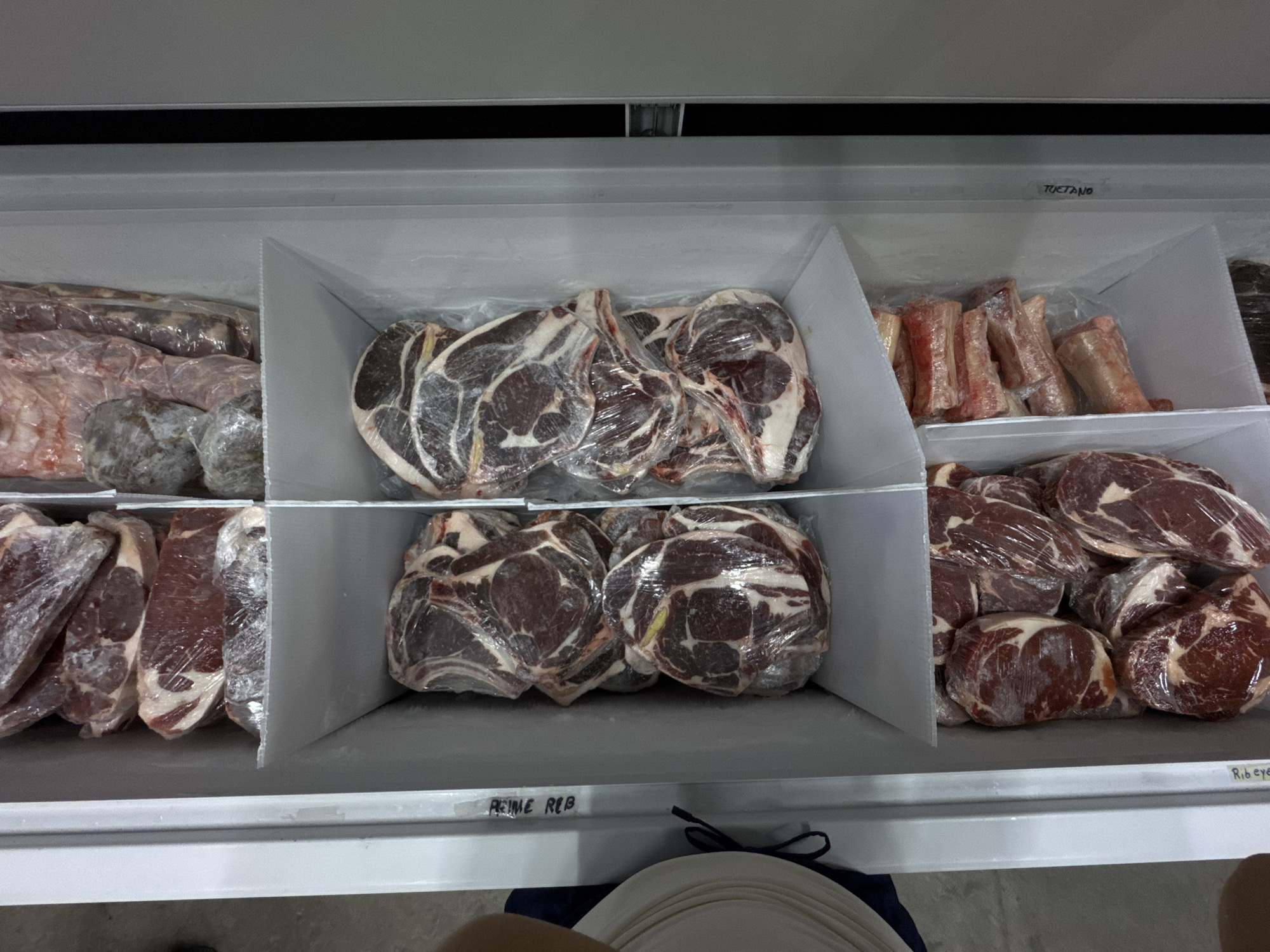A refrigerated display case contains various cuts of meat wrapped in clear plastic. The cuts are labeled "TOETANO," "REIME RIB," and "Rib eye." The location is likely a butcher shop or grocery store in Puebla, Mexico.