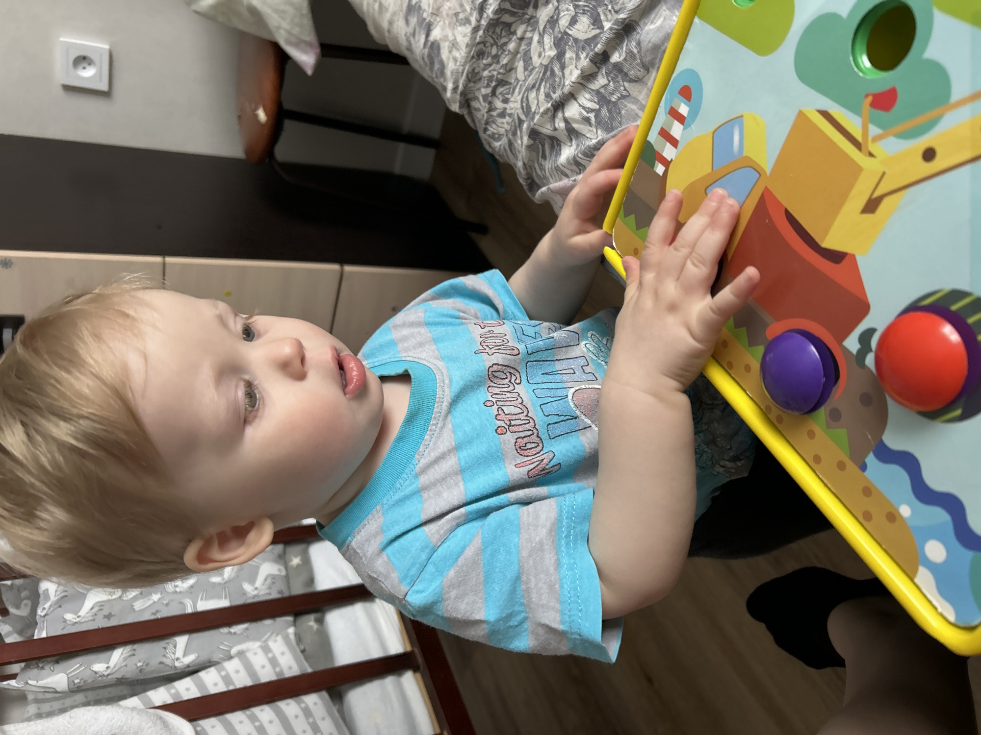 A young child, likely a toddler with fair skin, light blonde hair, and light-colored eyes, is actively engaged in playing with a vibrant activity board in an indoor setting. The child wears a blue and grey striped short-sleeved t-shirt, on which parts of the word "Waiting" and other graphics are visible. Both of the child's hands are on the colorful toy, which features yellow trim and illustrations of construction vehicles like a crane and an excavator, as well as a lighthouse. Interactive elements, including purple and red buttons, are visible on the board. The child's gaze is fixed on the toy, conveying a sense of focus and curiosity, with their mouth slightly ajar.
The background reveals a domestic environment, possibly a child's room or play area in Chaikovsky, Russia. To the left, part of a wooden crib or playpen with grey and white patterned bedding (some featuring animal designs) can be seen. Above the child's head, a white European-style electrical outlet is mounted on a light-colored wall, alongside dark and light-toned furniture. A bed with a grey and white floral-patterned comforter is partially visible in the upper background. The lighting suggests it is daytime. A dark shoe or foot is visible in the bottom right corner of the frame.