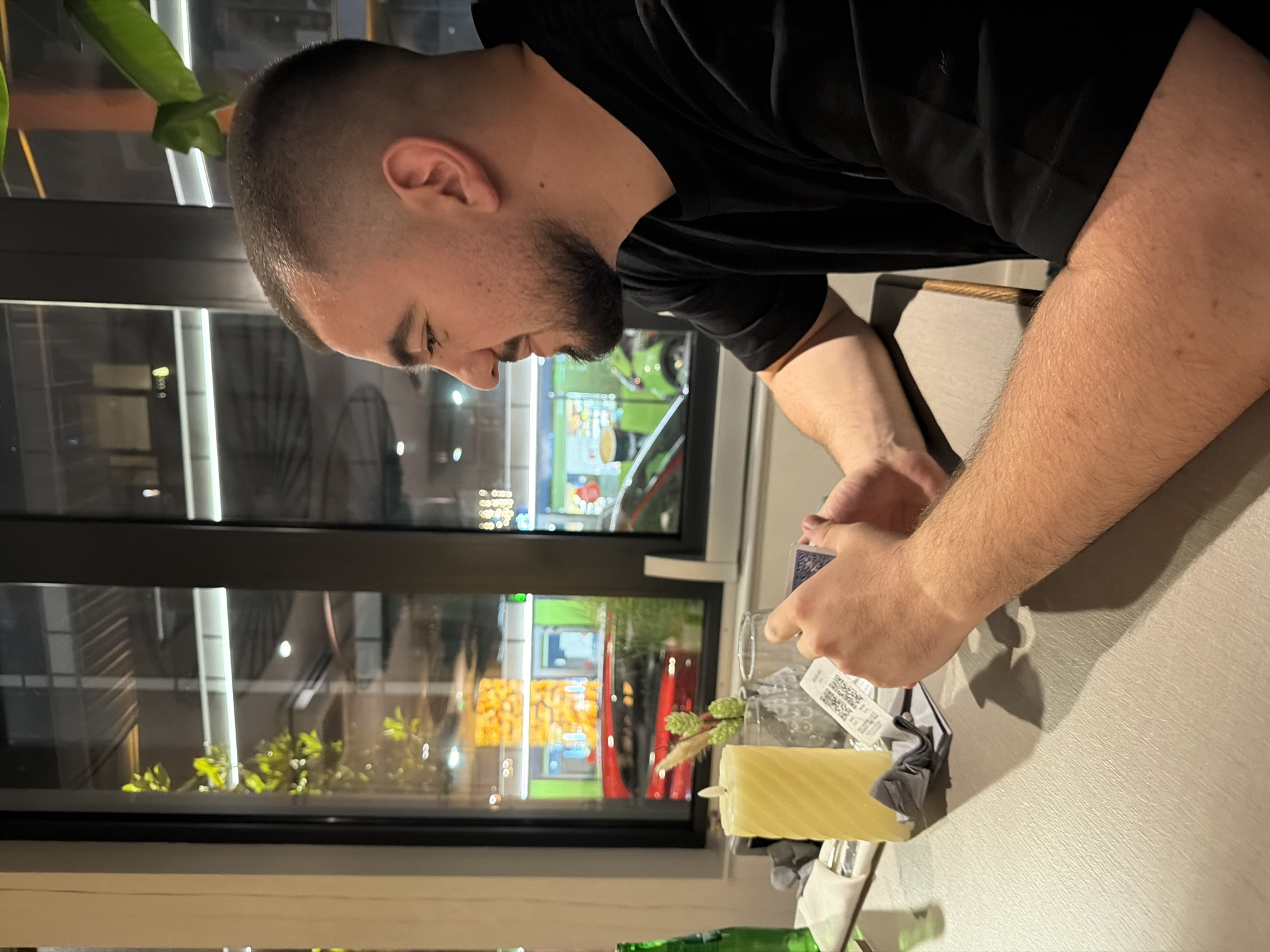 The image shows a man seated at a table next to a large window in what appears to be an indoor setting, likely a restaurant or cafe, in Sofia, Bulgaria. The man, seen from the chest up in profile, has a shaved head with a short beard and is wearing a black short-sleeved shirt. He is focused on a blue deck of playing cards held in his hands, suggesting he is either playing, examining, or performing a card trick.
On the light grey table, an unlit, spiral-fluted, light yellow candle stands beside a clear glass. A small, decorative sprig with green and brown elements is placed near the candle, alongside a crumpled grey napkin. Part of a green bottle is visible in the lower left corner.
The large window behind the man reflects the artificial lights of an outdoor street scene, indicating it is evening or nighttime. These reflections show various bright colors, including green, yellow, and red, likely from storefronts or signs. Some outdoor greenery is also visible through the window. A large green plant leaf partially obscures the upper left portion of the image, near the man's head. While some small text and a QR code are visible on a label attached to the glass and on the card deck, they are not legible. The man appears calm and concentrated on the cards.