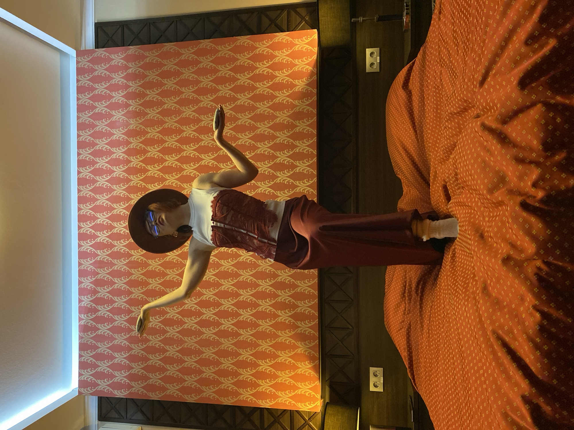 A person in a hat and a red outfit poses in a room. The room has patterned wallpaper in shades of orange and a bed with an orange patterned bedspread. The person is wearing a hat, blue sunglasses, a white top with a red corset, and red pants. The person's arms are raised in a dancing pose. The image could be taken at the Hotel Hyatt Regency Ekaterinburg in Russia.