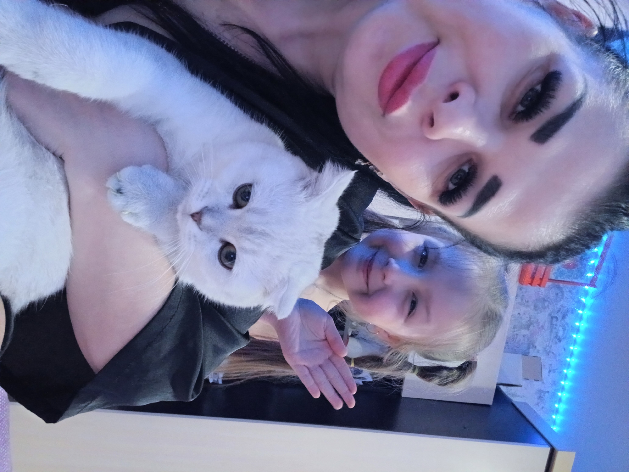 A selfie taken at close range features an adult female and a young girl, along with a white cat. The adult female has dark hair, heavy makeup, and is positioned at the top of the frame. She holds the cat, which appears to be looking at the camera. The young girl, with blonde hair styled in pigtails, is positioned lower in the frame and smiles towards the camera. They are likely indoors, with a blue light strip and what appears to be a wall visible in the background. The location is presumed to be Noviy, Russia.