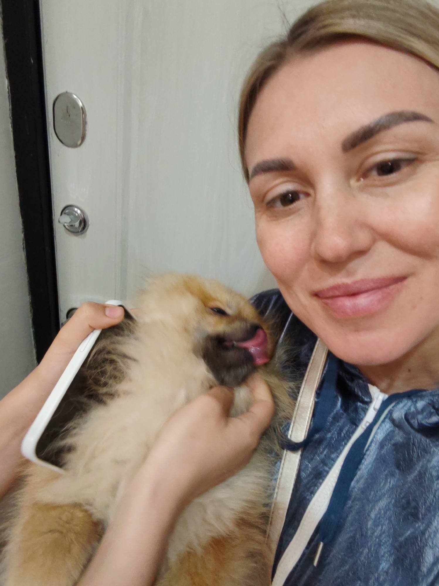 A woman is holding a fluffy Pomeranian puppy in her arms and posing for a selfie. The puppy is light brown and tan with its tongue sticking out, appearing to be licking the woman's hand. The woman has blonde hair pulled back and is smiling at the camera. She is wearing a blue, slightly shiny jacket with white and navy trim. The background appears to be a white door with two metallic door lock mechanisms, one oval and one circular. The lighting suggests it might be daytime. This image was taken in Komsomolsk-on-Amur, Russia.