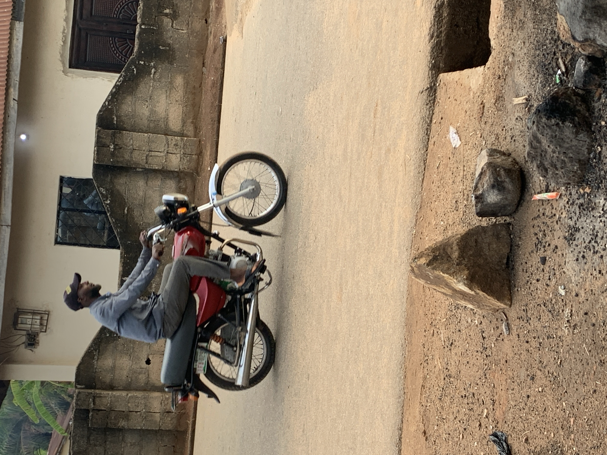 Akwanga, Nigeria: A man on a red motorcycle performs a wheelie on an asphalt road. He wears a gray shirt and pants with a baseball cap. The foreground shows a dirt shoulder with rocks and debris. A building with two windows is visible in the background.