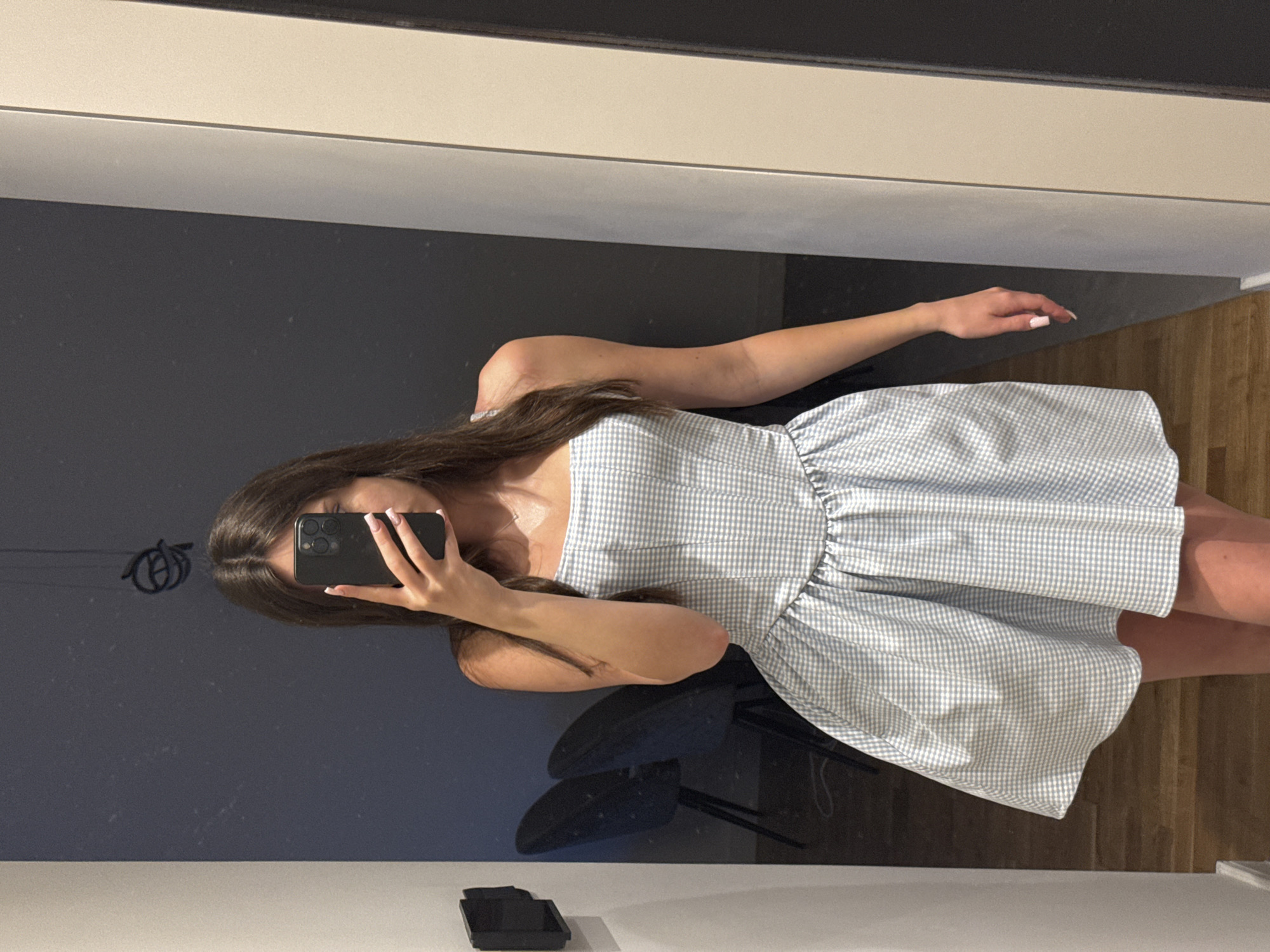 A young woman is captured in a mirror selfie, standing indoors. Her face is obscured by a black smartphone held in her right hand. She has long, dark hair that cascades over her shoulders. She is wearing a light blue and white gingham-patterned dress, featuring a fitted, possibly strapless or thin-strapped bodice and a full, gathered skirt that falls to her mid-thigh. Her left arm is bent slightly, and her fingers, adorned with pink-tipped manicured nails, are visible. A delicate necklace is visible around her neck.
The reflection in the mirror reveals a dark gray wall behind her and a light-colored wooden floor. Below the mirror's edge, in the foreground, there is a white surface with a small black object resting on it, possibly a phone accessory or a small container. The lighting is soft and even, suggesting an indoor setting during daytime. No specific location cues or emotions are discernible, and there is no visible text.