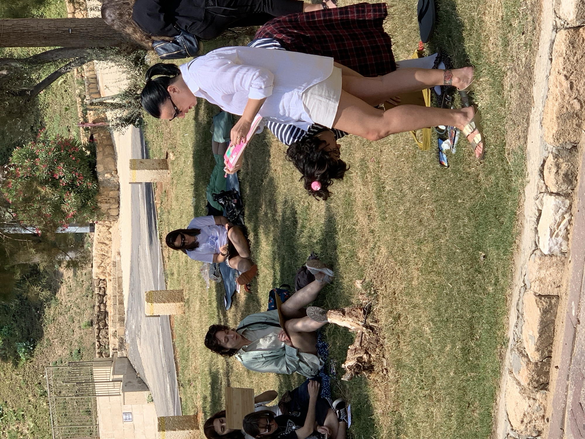 The image shows a group of people sitting on the grass in what appears to be a park. Two women are standing next to each other; one is wearing a white shirt and shorts, and the other is wearing a striped shirt. Other people are sitting on the grass, some with their legs crossed and others with their knees bent. There are trees and foliage in the background, as well as a paved path and a low stone wall. The photograph was taken in Tel Aviv, Israel.
