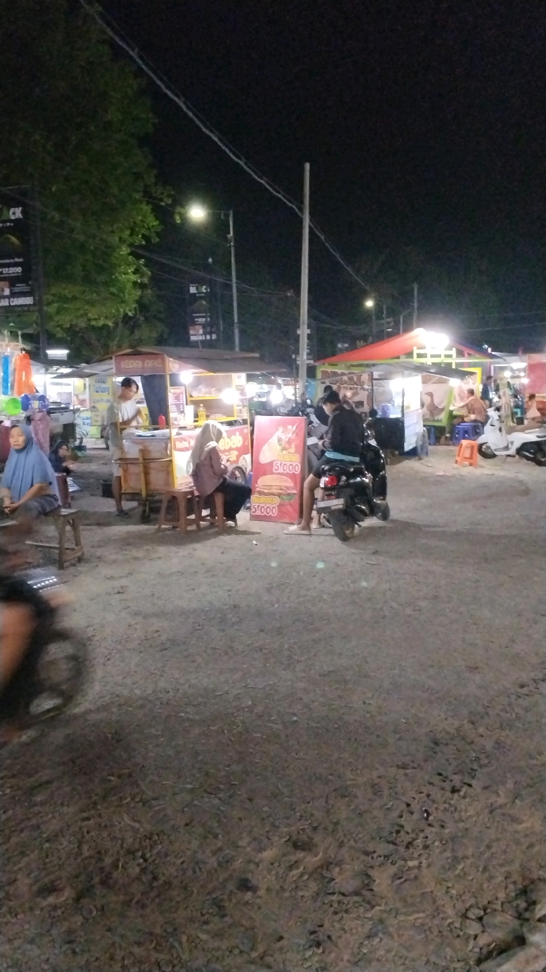 A night scene in Canggu, Indonesia, depicts a bustling street food market. The ground is uneven and covered in gravel and dirt. Several food stalls are visible, illuminated by bright lights, with awnings and tables set up. People are gathered around the stalls, some ordering food, others seated, and some standing. A prominent stall features a large sign with images of burgers and the text "Rp 51.000" displayed twice. Two individuals are seated on stools in front of this stall, while another person sits on a scooter. A black scooter is parked to the right of the stall, and a white scooter is parked further in the background. Trees with lush foliage line the left side of the scene, with streetlights and electrical wires overhead. Another stall in the background has colorful items hanging from it. The overall atmosphere suggests a vibrant and active evening market.