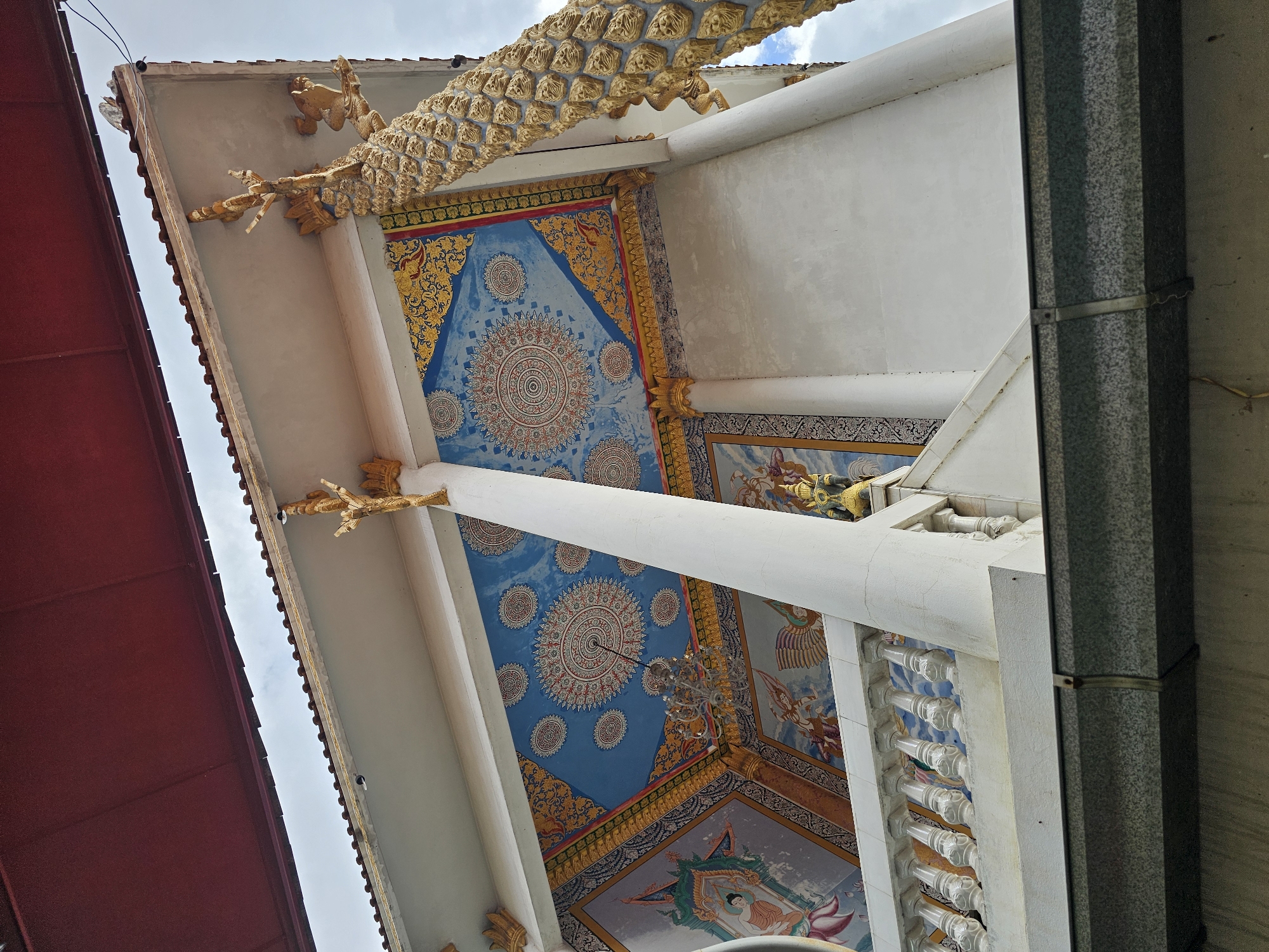 The image shows a low-angle view of the exterior of a Buddhist temple building in Bangkok, Thailand. The building features ornate white columns, a white railing on a raised platform, and a decorated ceiling with blue and gold motifs. A large, golden dragon-like sculpture adorns the corner of the building, its scales clearly visible. The ceiling is painted with circular patterns and images of figures. A chandelier hangs from the ceiling. Part of a red awning or roof is visible at the top of the image. The overall impression is one of intricate detail and vibrant color, typical of Thai temple architecture.