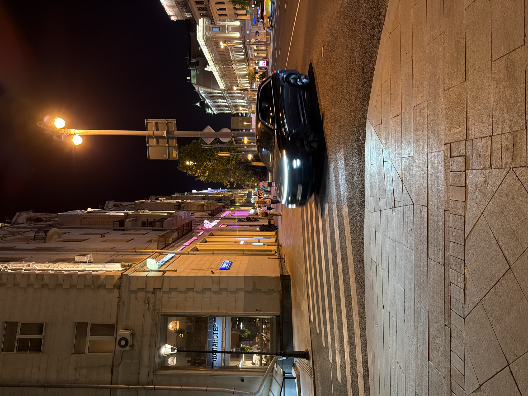The image depicts a nighttime street scene in Moscow, Russia. A dark-colored car is situated on the right side of the roadway, its headlights illuminated. Pedestrians can be seen crossing the street at a crosswalk. The buildings lining the street are multi-story and illuminated, displaying architectural details typical of Moscow. Streetlights are interspersed along the sidewalk. A street sign with Cyrillic characters is visible on one of the buildings. The pedestrian area is paved with light-colored stone.