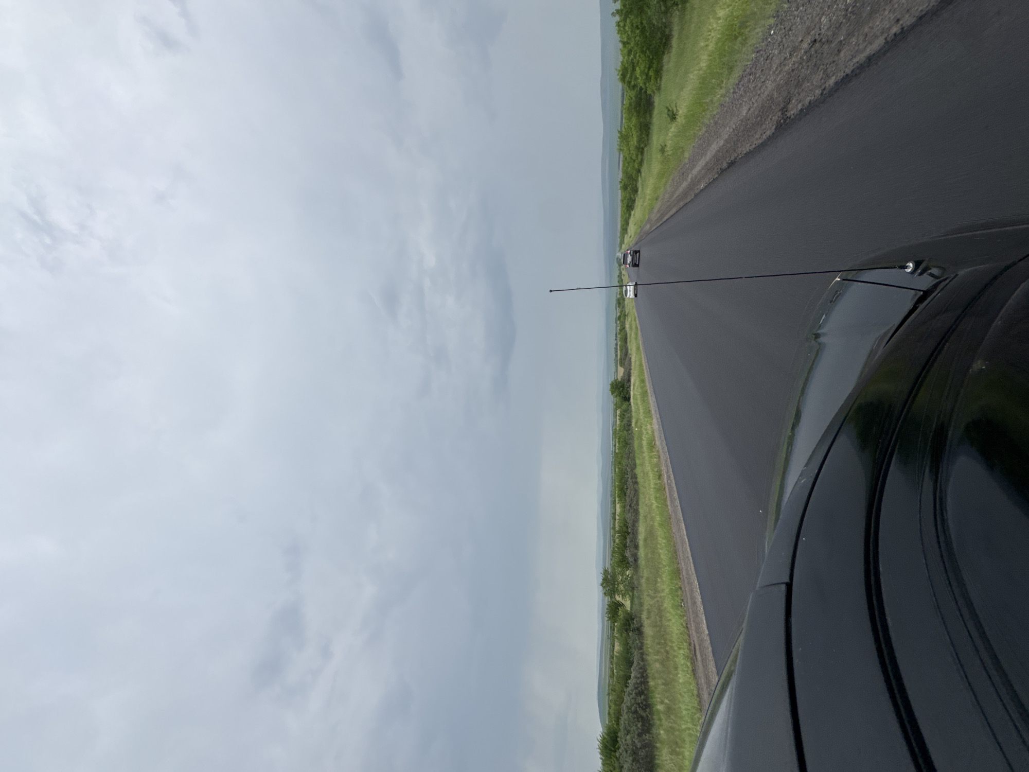 The image shows a view from the front of a black car driving on a paved road in an overcast, rural landscape. The road stretches into the distance, with green fields and low, rolling hills on either side. A few other vehicles, a white car and a dark-colored SUV, are visible ahead on the road. The sky is filled with heavy, gray clouds. The car's hood and a tall antenna are in the foreground, indicating the perspective of the driver or passenger. The location is identified as the city of Сарагаш, Russia.