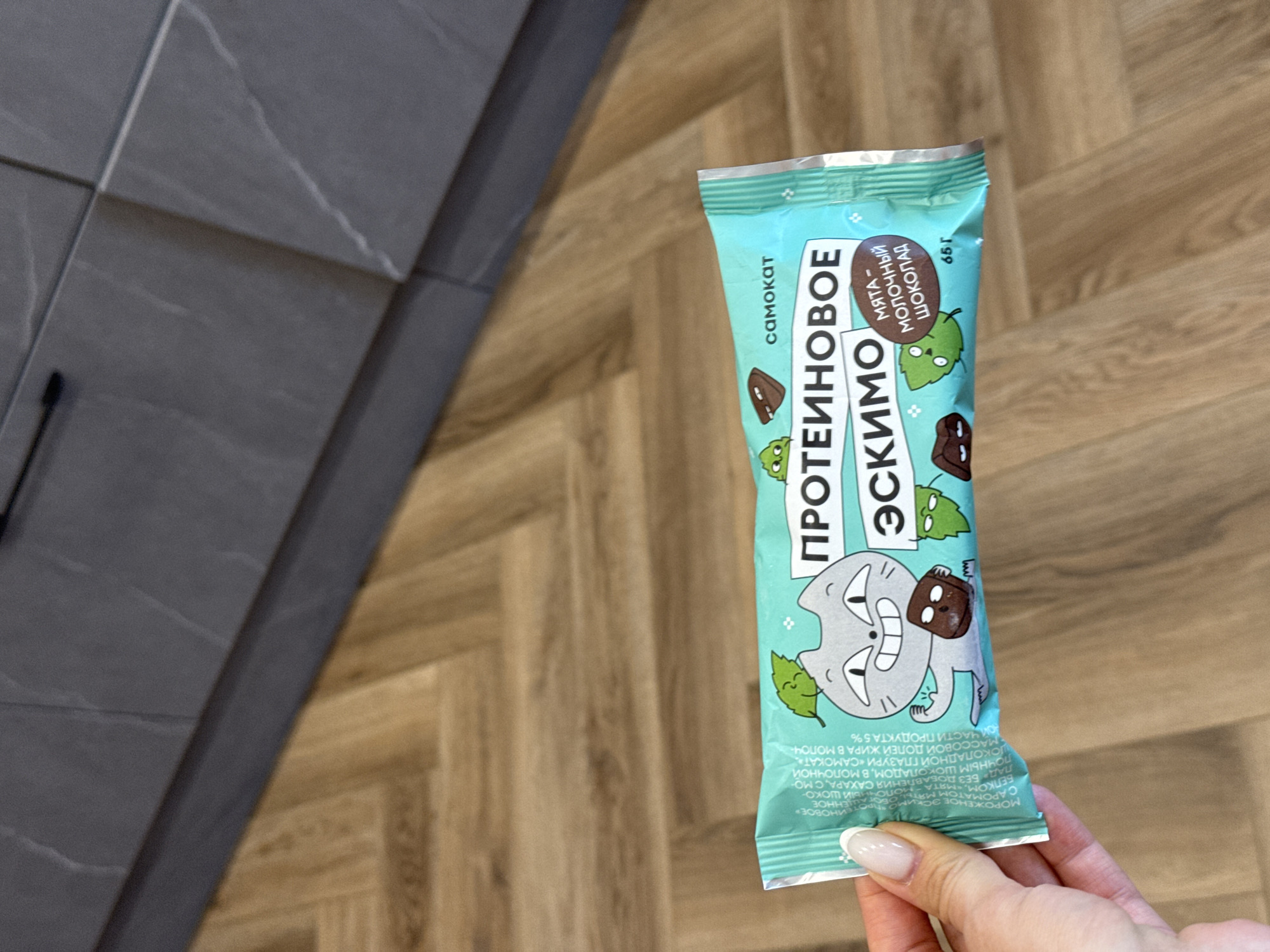 An eye-level medium shot shows a hand holding a light blue ice cream bar wrapper in front of a wood-paneled floor and dark gray cabinetry. The wrapper is adorned with a cartoon cat holding a chocolate square and a text that reads "ПРОТЕИНОВОЕ ЭСКИМО" (Protein Eskimo) in large font. The text "МЯТА-МОЛОЧНЫЙ ШОКОЛАД" (Mint-Milk Chocolate) is in a smaller oval shape on the right side of the wrapper. The ice cream bar appears to be a product of "САМОКАТ" (Samokat), a Russian food delivery service. The hand holding the wrapper has manicured nails with a French tip design. The background is a herringbone pattern of light brown wood flooring and dark gray kitchen cabinets with subtle marble-like veining. The lighting is bright and diffused, suggesting an indoor setting.