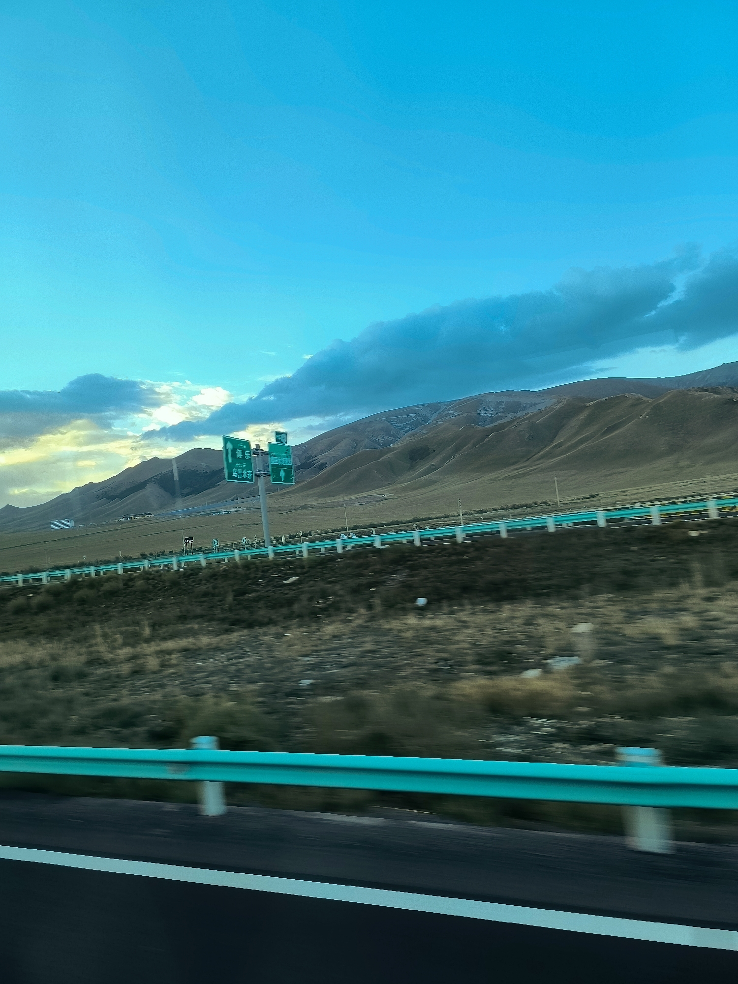 An eye-level, medium shot captures a vast, open landscape with a focus on a highway. The sky above is a vibrant, almost artificial-looking turquoise, with wispy clouds scattered across it. To the right, darker, more dramatic clouds create a striking contrast against the bright sky. Rolling hills, covered in dry, golden-brown vegetation, dominate the middle ground. A highway stretches horizontally across the scene, separated by a bright turquoise guardrail with white posts. In the distance, several green highway signs are visible, indicating directions with Chinese characters. The foreground shows a blur of asphalt and a white lane marker, suggesting the viewer is in a moving vehicle. The overall impression is one of a vast, somewhat desolate, and remote highway environment in China, likely in a mountainous region.