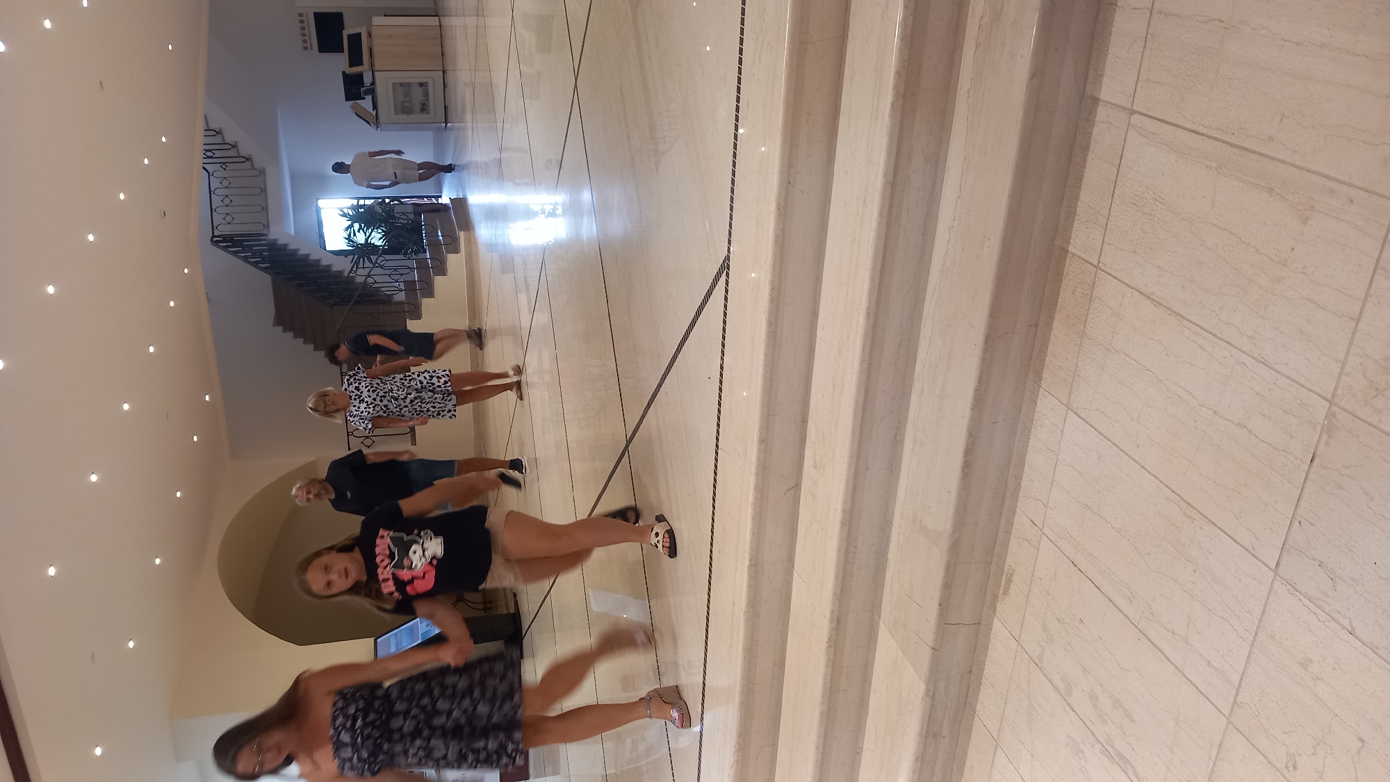 The image depicts the interior of a hotel lobby in Kos, Greece, featuring a group of people walking on a polished marble floor. In the foreground, a woman in a dark strapless dress is walking towards the left, holding hands with a young girl wearing a black t-shirt with a graphic print and light-colored shorts. Behind them, a man with gray hair and a dark t-shirt walks towards the right. Further back, a woman in a black and white patterned dress is seen, and behind her, a man in a dark t-shirt and shorts is walking down a staircase. A man in a white t-shirt and shorts is visible walking away from the camera on the right side of the frame. The lobby has an arched ceiling with recessed lighting, and a curved staircase with a decorative railing is prominent in the background. The floor is a light-colored marble with dark inlay lines. The environment appears to be a modern hotel or resort.
