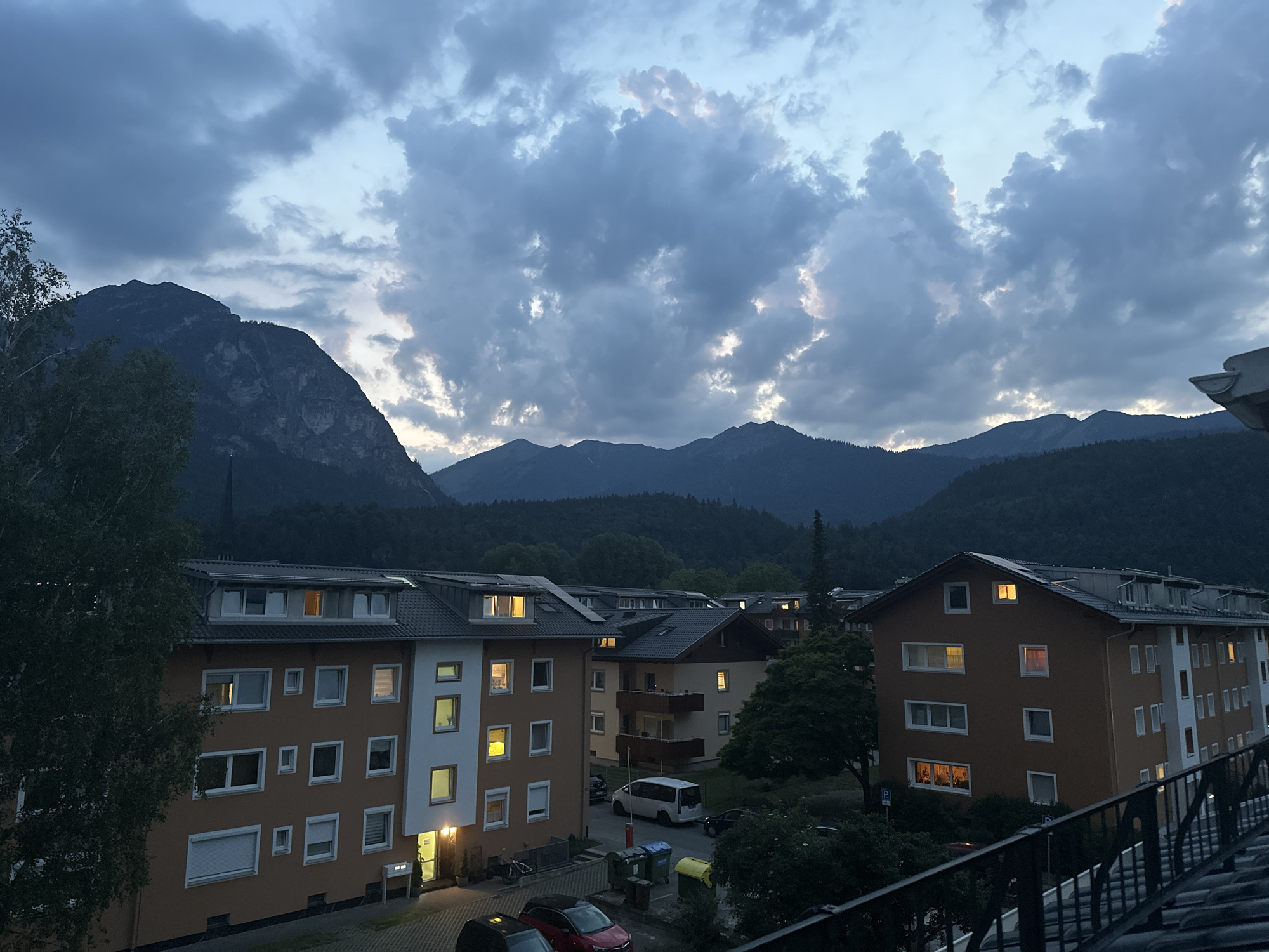 The image depicts a view of apartment buildings in Garmisch-Partenkirchen, Germany, at dusk. In the foreground, apartment buildings with illuminated windows are visible. A large tree stands to the left, obscuring part of one building. In the middle ground, several more apartment buildings and parked cars are present, along with trash receptacles. The background features a dense forest that transitions into mountainous terrain under a cloudy sky. The overall lighting suggests it is either early morning or late evening.
