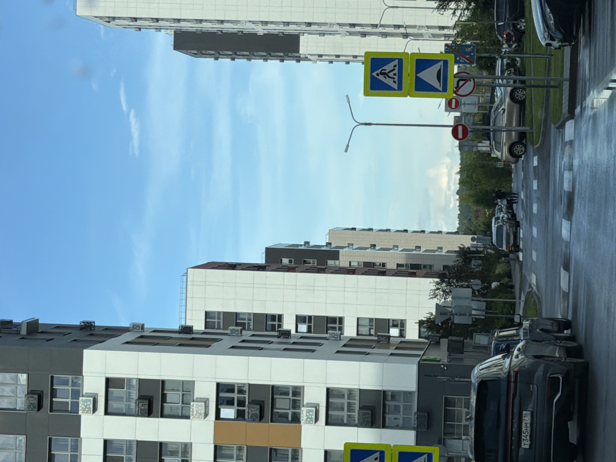 The image depicts a street scene in a modern urban environment, likely in Kazan, Russia. Two high-rise apartment buildings dominate the left and right sides of the frame, characterized by their contemporary architectural style with segmented facades in white, grey, and some brown accents. The buildings feature numerous windows and visible external air conditioning units. A bright blue sky with scattered white clouds forms the backdrop.
In the foreground, a street is visible with white pedestrian crossing markings. Several vehicles are present, including a dark SUV with a license plate that reads "T345HM 716", and other cars parked or driving on the road. Traffic signs are also prominent, including pedestrian crossing signs (a blue square with a white pedestrian silhouette) and a speed bump warning sign (a blue square with a white inverted triangle and three humps). Other signs with red circles, indicating prohibitions, are also visible.
A lamppost with two lights stands in the middle ground. On the right side of the street, trees and greenery are present, suggesting a landscaped urban setting. The street appears wet, indicating recent rain. A few pedestrians are visible in the distance near the buildings on the left. The overall impression is a well-developed residential area with modern infrastructure.