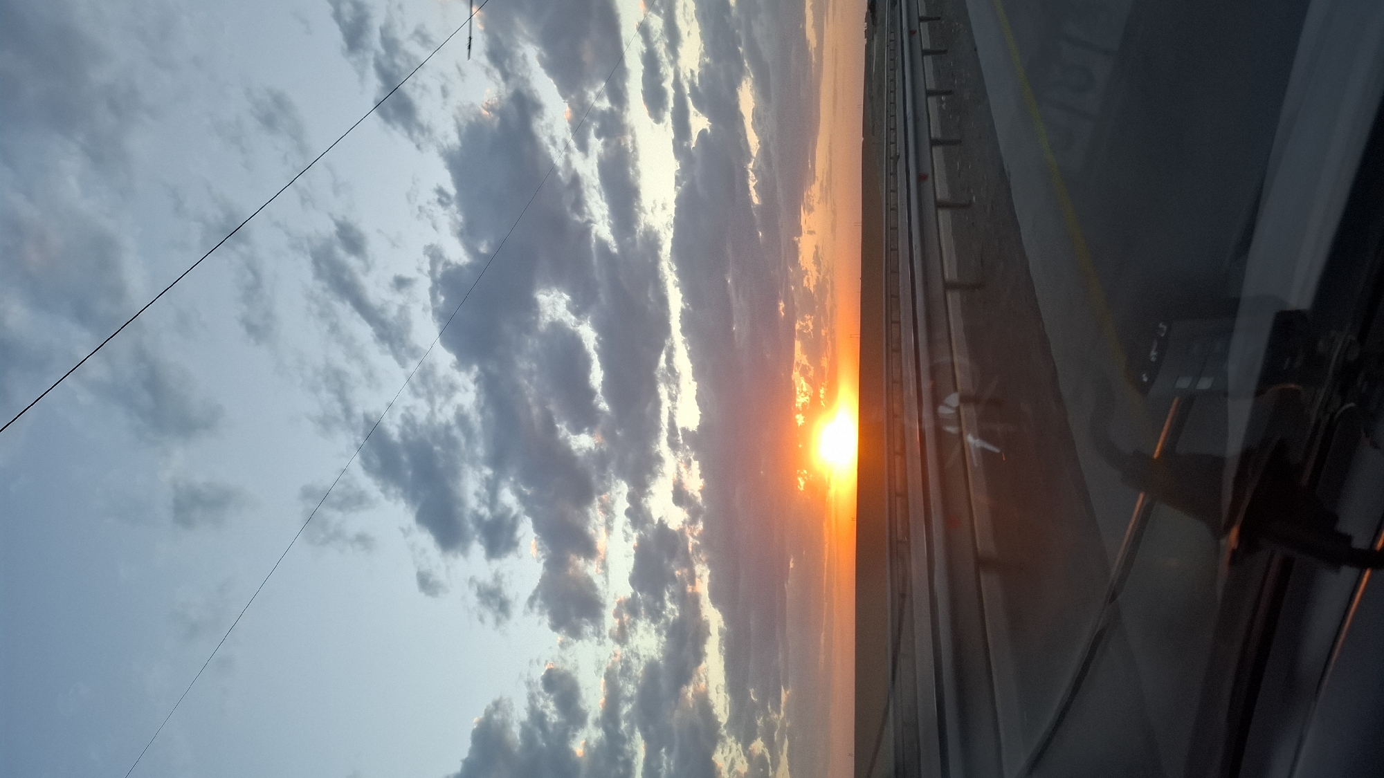 The image captures a sunset viewed from inside a vehicle, likely on a highway in or near Shymkent, Kazakhstan. The sun is a bright orange orb positioned low on the horizon, partially obscured by clouds that are tinged with pink and orange. The sky transitions from a pale blue near the top to warmer tones as it approaches the horizon. A dark, flat landscape stretches across the middle ground, with a highway barrier visible in the foreground, separating the road from the undeveloped land. The interior of the vehicle is reflected in the windshield, showing parts of the dashboard and wiper blades. Two dark, thin lines, possibly power lines or wires, stretch diagonally across the upper portion of the sky. The overall impression is one of a scenic drive at dusk.