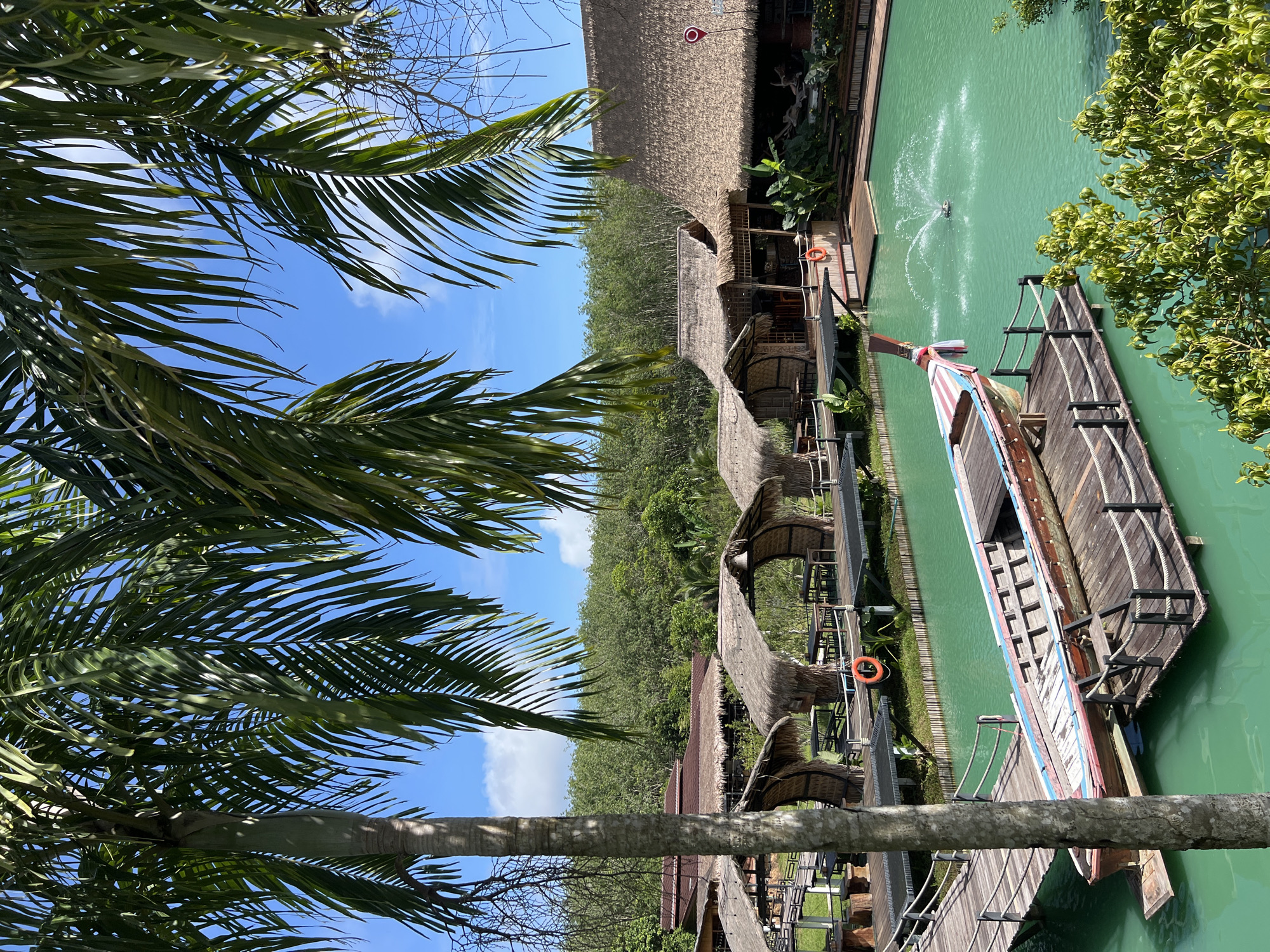 This media file depicts a scenic waterside resort or restaurant in Tambon Pa Kae Bo Hin, Thailand. The foreground is dominated by the lush green fronds of a palm tree, framing a view of the tropical setting. The scene is bathed in bright sunlight, suggesting it's daytime.
The central focus is a vibrant turquoise body of water, likely a river or canal, where a long, narrow wooden boat with a red, white, and blue striped trim is docked. The boat has a traditional design and appears to be a prominent feature of the location. In the water, a fountain or water jet creates a dynamic spray.
Along the water's edge, a series of thatched-roof structures, possibly huts or dining areas, are connected by wooden walkways. These structures have a rustic, natural aesthetic. Orange life preservers are visible, indicating a focus on water safety. Beyond the resort area, a dense forest of green trees stretches towards the horizon under a clear blue sky with scattered white clouds.
The overall impression is one of tranquility and natural beauty, characteristic of a tropical vacation destination. No people are explicitly visible engaging in activities, but the presence of dining areas and the boat suggests leisure and potential tourism. The time of day is clearly daylight, and the weather appears to be sunny and pleasant. The location cues point to a tropical environment, likely Southeast Asia, specifically Thailand as indicated by the location context.