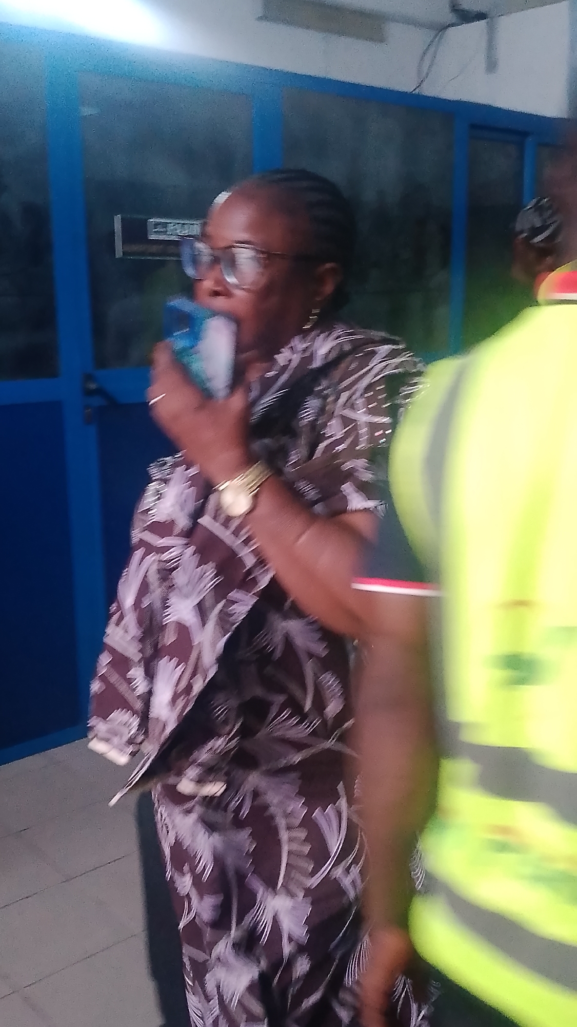 The image shows two individuals in an indoor setting, possibly an office or a hallway with blue-framed glass doors in the background. The primary subject is a woman in the foreground, wearing glasses and a patterned dress. She is holding a blue object to her mouth, and her expression suggests she might be speaking or eating. A gold watch is visible on her left wrist. To the right of the woman, another person is partially visible, wearing a bright yellow vest, suggesting they might be involved in some official capacity or work-related activity. The lighting is dim, hinting at an indoor environment at an unspecified time of day. The overall scene appears to be a casual or semi-formal interaction. No visible text is decipherable in the image.