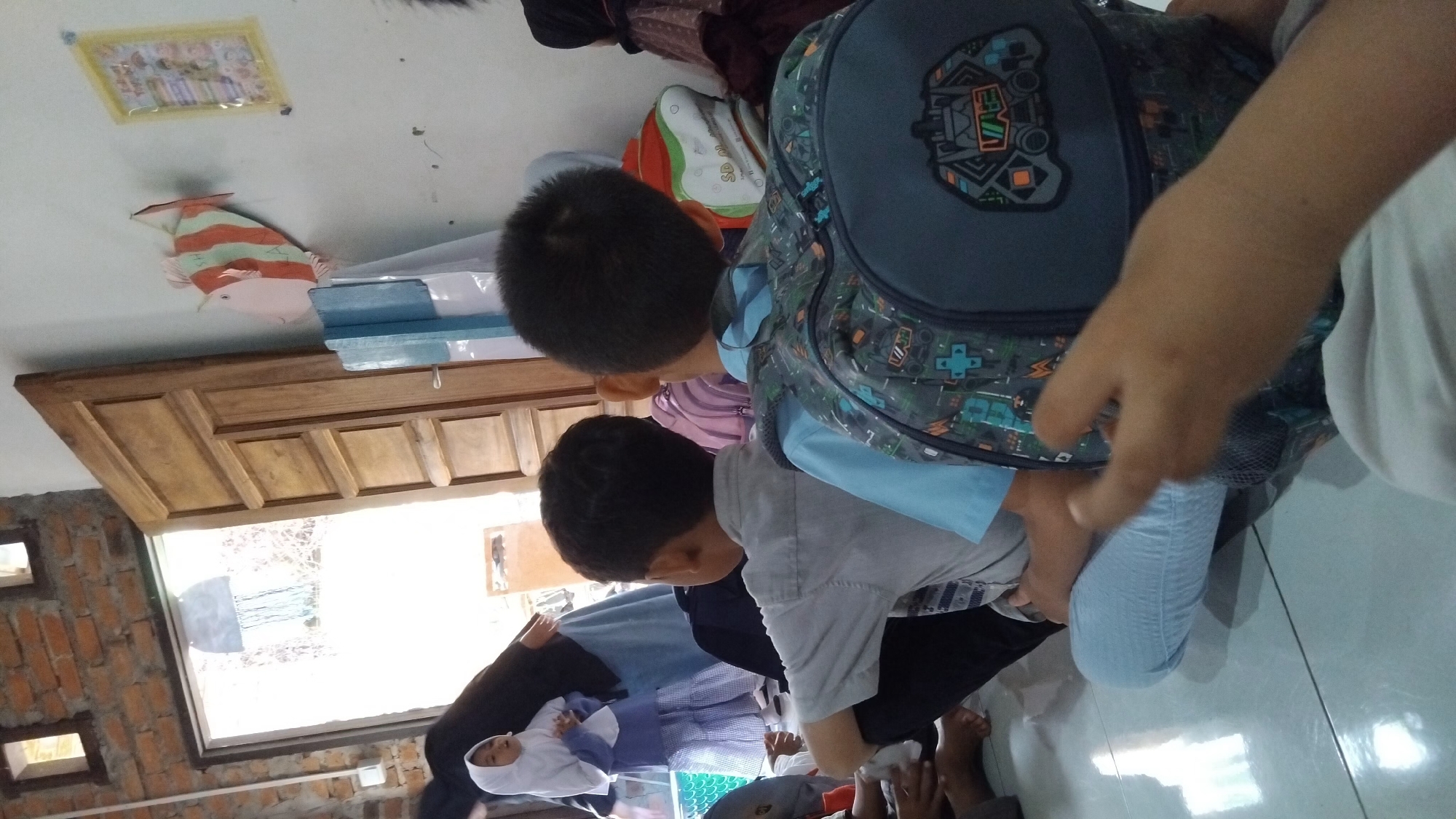 The image depicts a group of children sitting on a tiled floor, likely indoors, with a wooden door and a brick wall in the background. The children are facing away from the camera, with their backs to the viewer. One child in the foreground is wearing a light blue shirt and a patterned backpack. Another child next to them is wearing a light grey shirt. A younger child in a white hijab and blue dress is visible to the left, standing near an adult dressed in dark clothing. Decorations, including a paper fish and a framed picture, are visible on the wall. The lighting suggests an indoor environment, possibly a classroom or a home. The location is specified as Bangun Rejo, Indonesia.