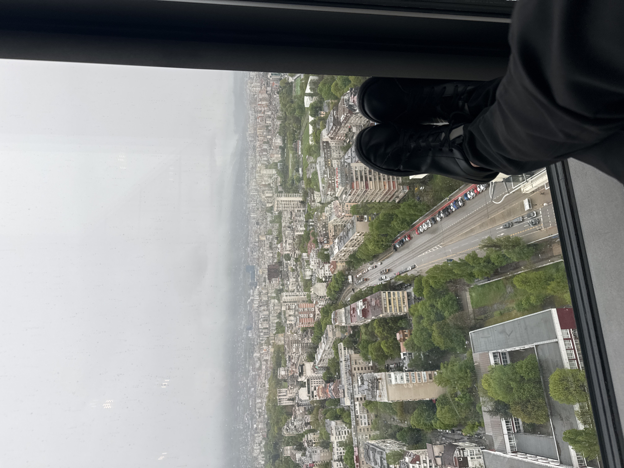 The image captures an expansive urban landscape as seen from a high vantage point, looking down through a window. In the foreground, positioned at the bottom right, are the legs and feet of a person wearing black shoes and dark trousers. Their feet appear to be casually resting near the window frame, suggesting a relaxed observation of the view.
The scene unfolds into a sprawling city, identifiable as Sofia, Bulgaria. The sky on the left side of the frame is uniformly overcast and gray, indicating a cloudy day. Visible water droplets or smudges on the window glass suggest recent rain or condensation.
Below, a wide multi-lane road or highway stretches across the middle ground, populated with cars and other vehicles. Along one side of this road, a prominent row of parked cars, distinctively red and white, creates a visible line. The urban environment features a dense array of buildings, varying in height and architectural styles, interspersed with numerous green spaces, parks, and trees. The city extends towards a hazy horizon, where structures become less distinct. No discernible text is visible within the image.