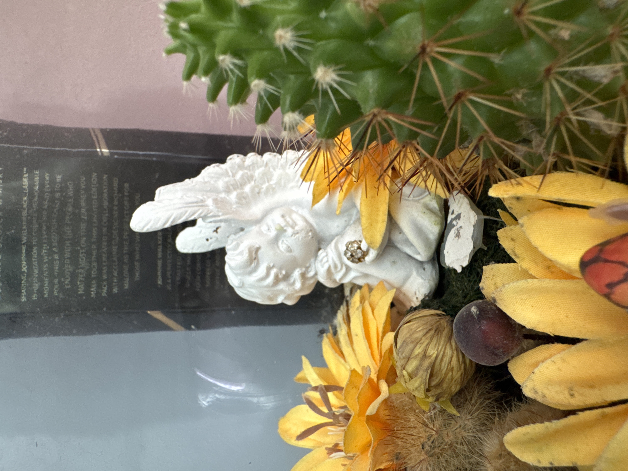 The image presents a close-up view of a decorative arrangement. A white, possibly plaster or ceramic, cherub statue is nestled amongst a prickly green cactus, yellow artificial flowers, and dried floral elements. The cherub is adorned with a decorative jewel. A portion of a black box labeled "SHARING JOHNNIE WALKER BLACK LABEL..." is visible, providing text details. The box inscription mentions an invitation to create memorable moments, collaboration with designer Mohard Malone, and a limited edition pack. No geographical information is provided, so it's not possible to confirm the location as Burgas, Bulgaria.