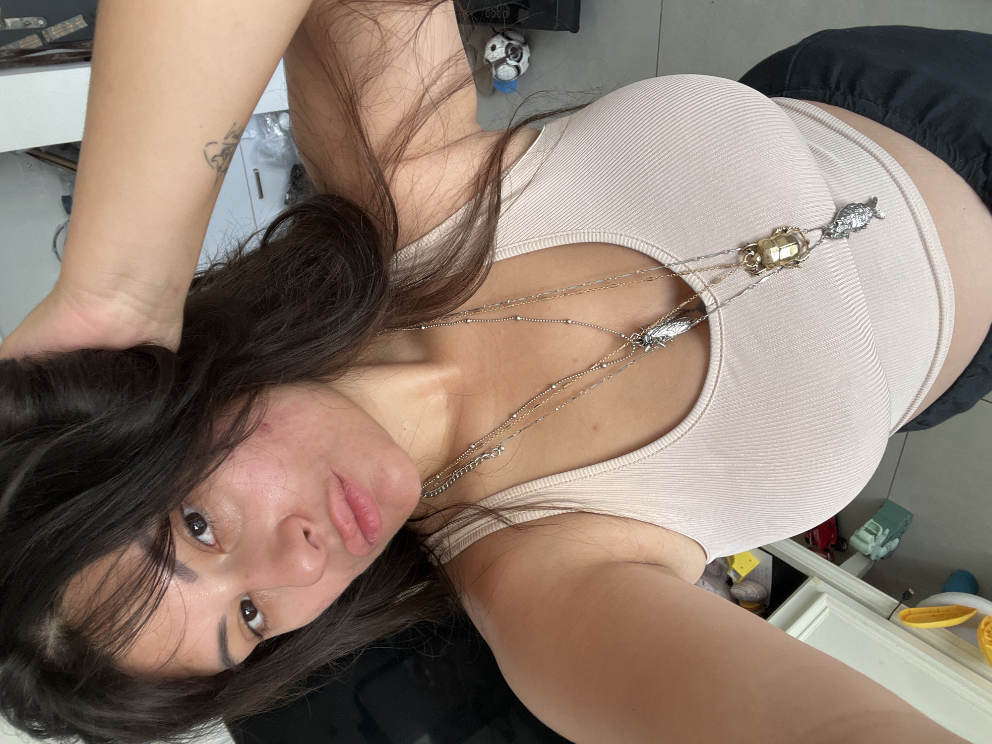 The image shows a young woman with long, dark hair and brown eyes, pouting her lips. She is wearing a beige ribbed tank top and several layered necklaces, including pendants shaped like insects and a fish. A small tattoo is visible on her left forearm. The background suggests an indoor setting with furniture and some toys on the floor, indicative of a home environment. The lighting appears to be natural, suggesting daytime.
