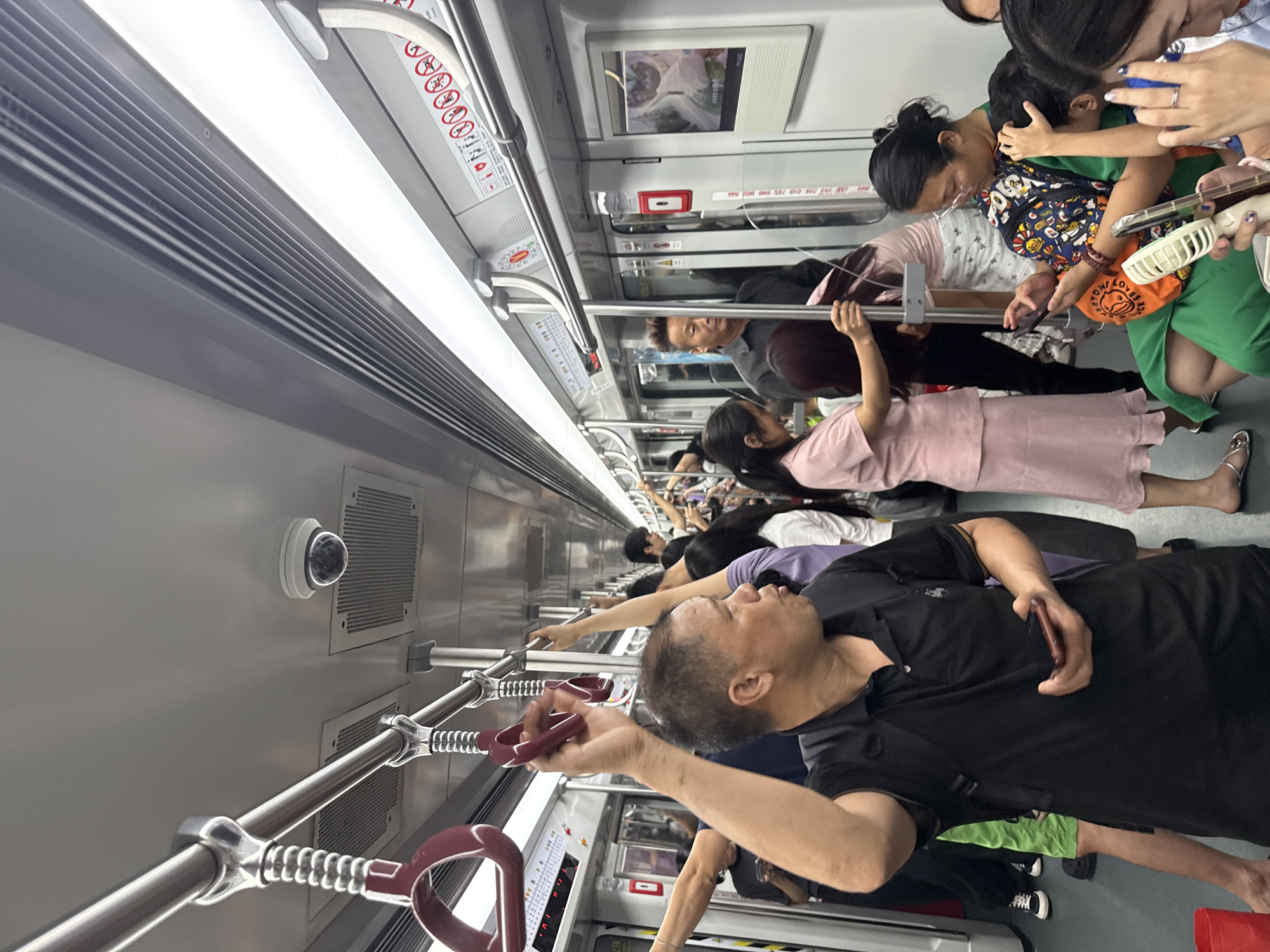 The media file depicts a crowded subway car in Haizhu District, China. The subjects are numerous passengers, including adults and children, in various states of standing and sitting. They are surrounded by typical subway interior elements: silver grab bars, ventilation grilles, lighting fixtures, and advertisements on the walls. The scene is brightly lit by the interior lights, suggesting it is daytime.
The primary activity is commuting, with passengers holding onto poles for stability, engaging with their phones, or interacting with each other. One notable interaction involves a woman holding a child and another woman, possibly a child, embracing the child. Another passenger, a man in a black polo shirt, is looking towards the camera and holding a phone. The overall mood appears to be one of crowded normalcy, with individuals focused on their personal space or devices amidst the throng.
Specific objects of note include a security camera mounted on the wall, a screen displaying an image, and a red emergency call button. Some passengers are carrying bags, and one person is wearing a colorful printed vest. The presence of numerous people and the enclosed environment suggest a busy urban commute. The image provides a snapshot of daily life on public transportation in a densely populated area.