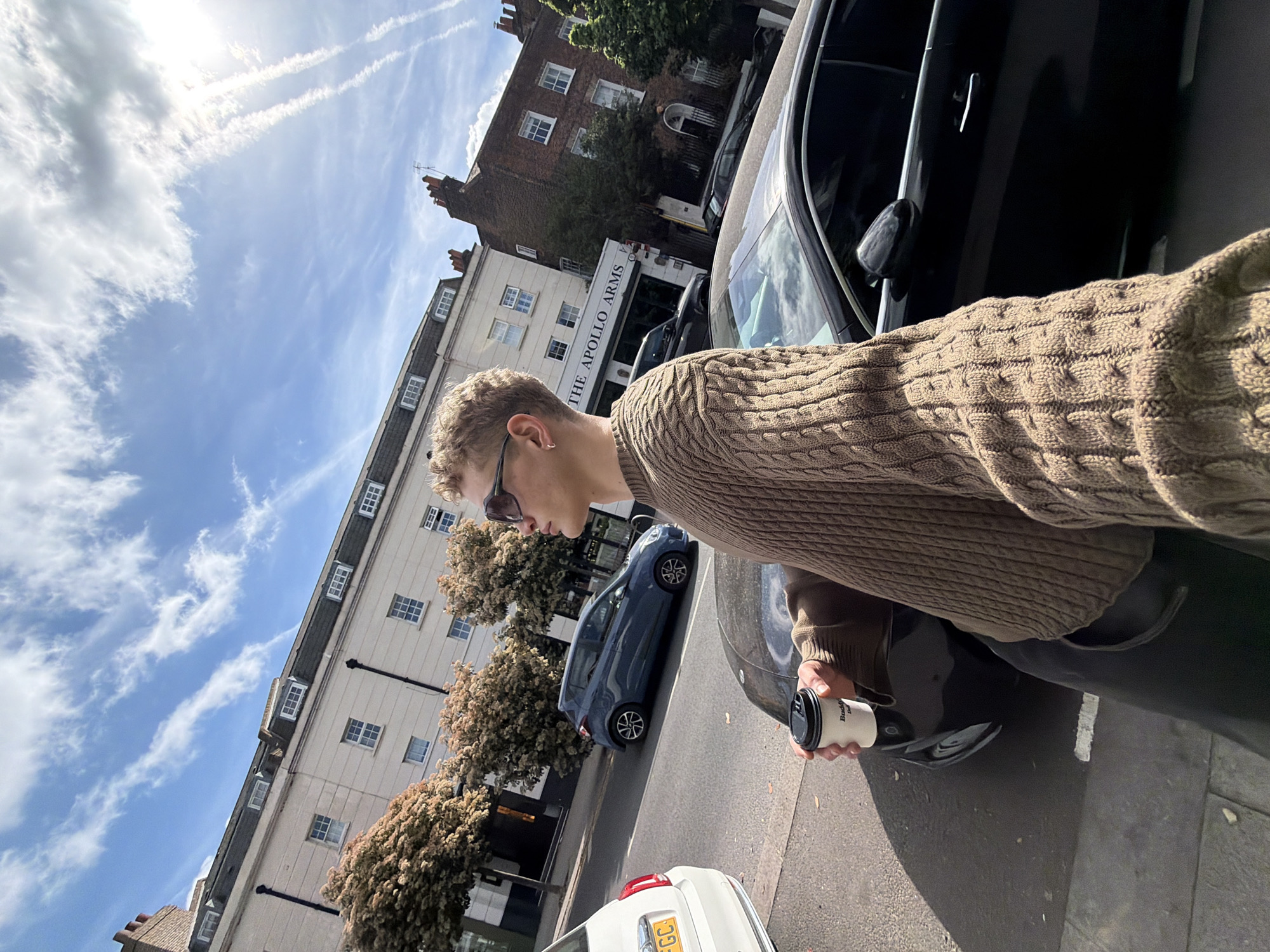 A medium shot captures a light-skinned male, with short blond hair and sunglasses, in a brown cable-knit sweater. He appears to be holding a coffee cup with a black lid in his right hand while standing next to a black car with an open door. In the background stands a building with white facade labeled "The Apollo Arms" with rows of windows. Beyond this building, another brick building is visible. A blue car and a white car are parked along the street. The sky is blue with some white clouds and visible contrails. The photograph suggests a street scene in London, United Kingdom, based on the architectural style and signage visible in the background.