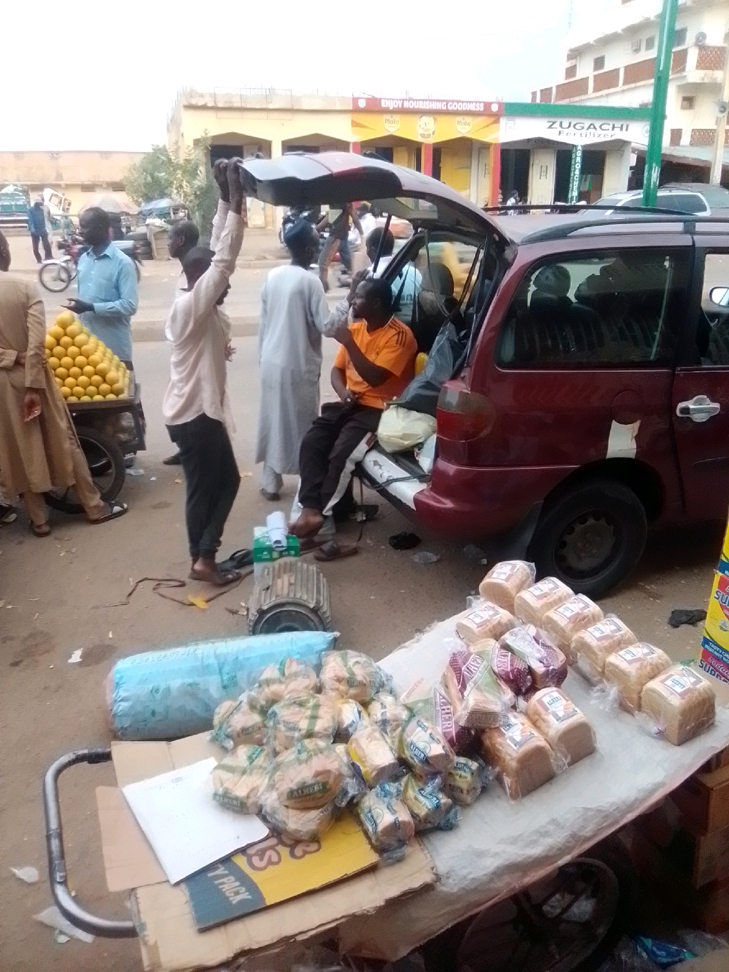 The image depicts a busy street scene in Arawa, Nigeria, with a focus on vendors and their goods. In the foreground, a cart is laden with numerous loaves of bread, some individually wrapped and others in larger plastic packaging. The bread is arranged on a white cloth covering the cart. To the left of the cart, a large blue tarp or sack is visible. Further back, a vendor in an orange shirt is seated next to a car with its trunk open. Another individual, holding a large object overhead, stands near a pile of yellow citrus fruits on a wheeled cart. Several other people are milling about in the background, some interacting with vendors. The environment appears to be an outdoor market or street-side shopping area, with buildings visible in the distance. The ground is paved and appears dusty, characteristic of a street. The overall impression is one of a lively commercial exchange.