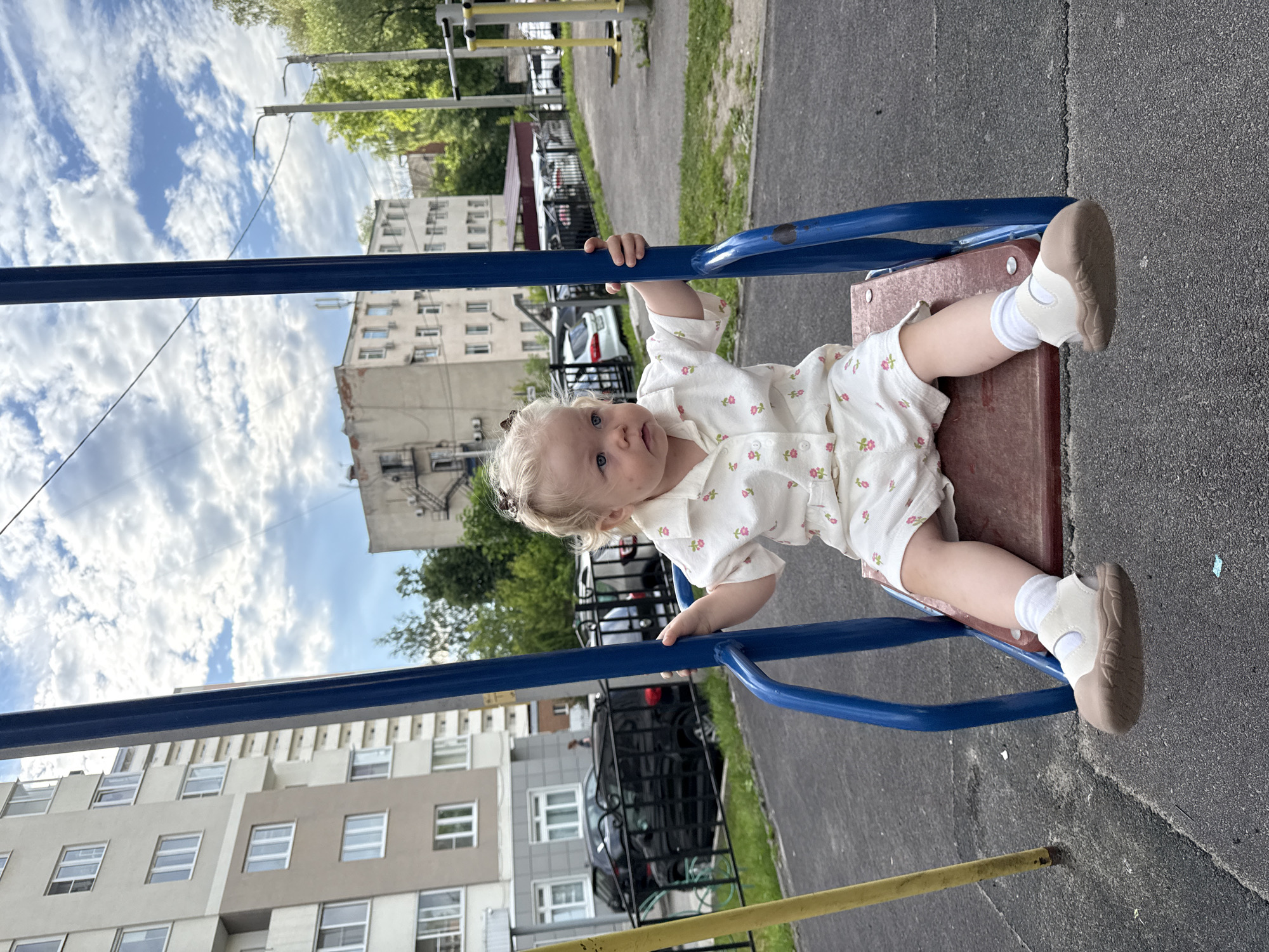 A blonde toddler is sitting on a swing in a playground. The toddler is wearing a white outfit with a floral print and white socks and sandals. The swing set is blue and red. In the background, there are apartment buildings, cars, trees, and a cloudy sky. The location is a playground in the city of Vladimir, Russia.