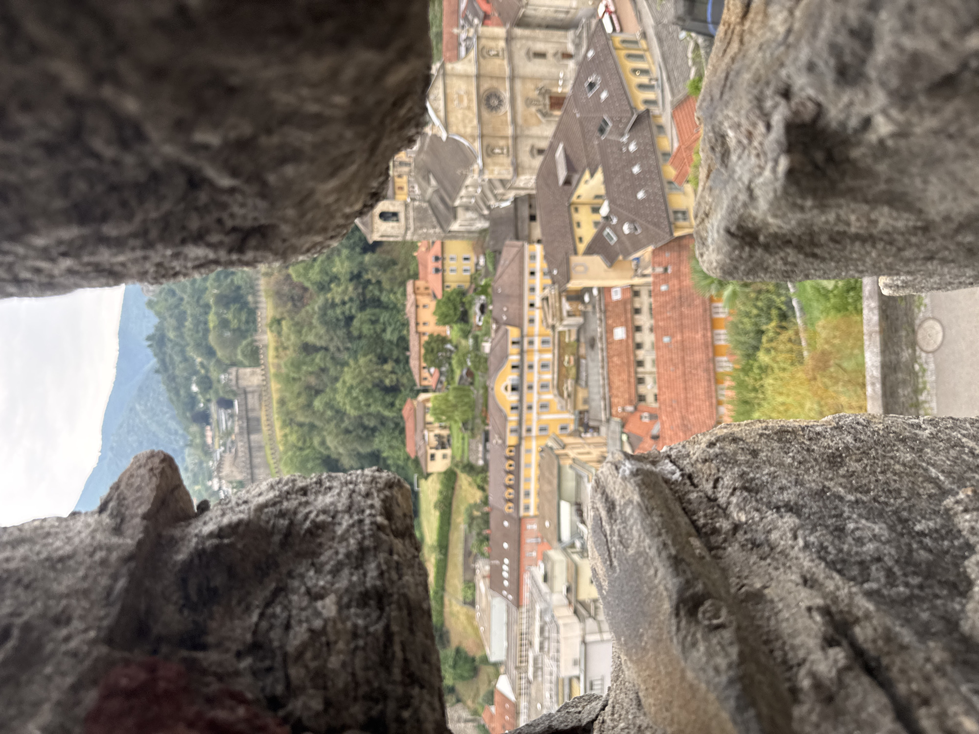 The image is a view through a gap in rough stone walls, looking out over the city of Bellinzona, Switzerland. The foreground is dominated by the textured, grey stone of the walls, which partially obscure the view. Beyond the walls, a sprawling cityscape unfolds, characterized by buildings with terracotta tiled roofs. Prominently featured is a large, ornate church with a bell tower, its facade rendered in a light stone color. Other buildings, varying in size and color, including some yellow structures, are clustered together. In the background, a fortified wall runs along a hillside covered in green trees, with further hills and mountains rising behind it under an overcast sky. The perspective suggests the viewer is elevated, looking down upon the city and its fortifications. There are no people or active interactions visible in the scene.