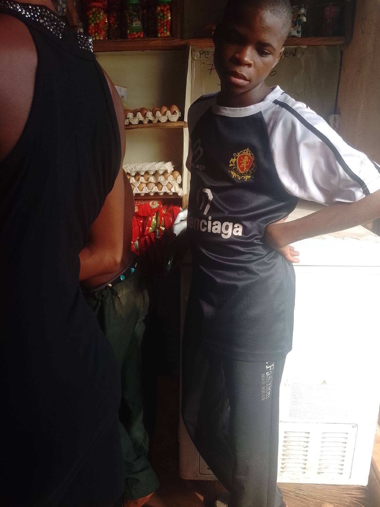 The image depicts a young male in Ejule, Nigeria, standing in front of a white refrigerator inside a small store. He is wearing a black and white soccer jersey with "Balenciaga" printed on it, and black sweatpants with the word "Fashion" printed vertically along the leg. He has his hand on his hip. A woman in a black dress stands partially in the foreground, obscuring the view of the products on the shelves, including eggs, snacks, and bottled goods. The store has basic wooden shelves stocked with various items for sale. A chalkboard can be seen partially in the background behind the shelves.