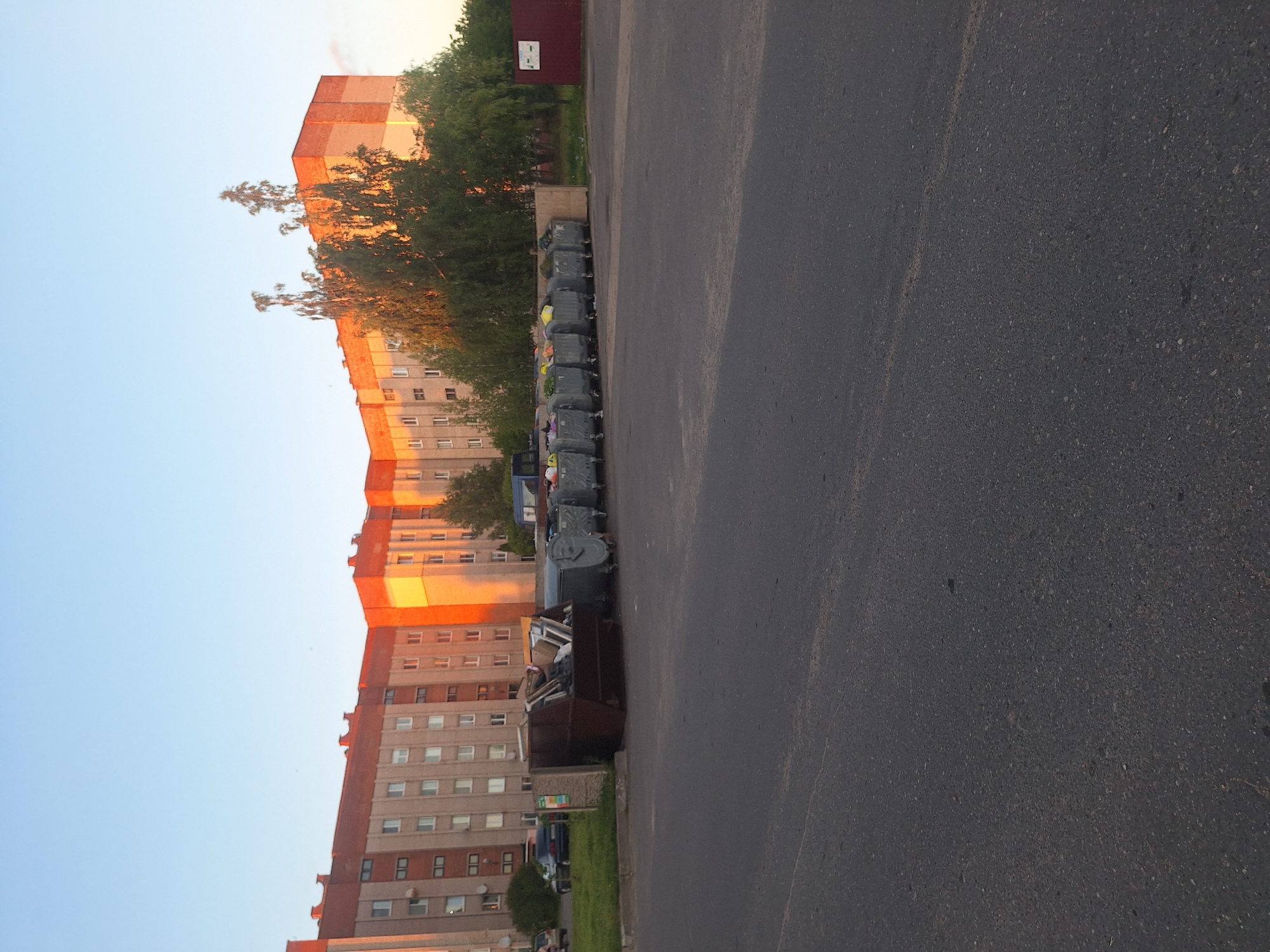 In Kapyl District, Belarus, a row of large, industrial-style dumpsters stands in a paved area adjacent to a grassy strip. The dumpsters are mostly filled with waste. Behind the dumpsters, a tall, multi-story residential building with a brick facade and numerous windows is visible. The building is illuminated by the warm, orange glow of the setting sun. Trees and foliage are present between the dumpsters and the building. In the foreground, a dark asphalt surface dominates the lower portion of the image, suggesting a road or parking lot. A dark-colored car is parked on the grass to the left. The sky is clear and shows the blue of daytime with the setting sun casting orange hues on the building.