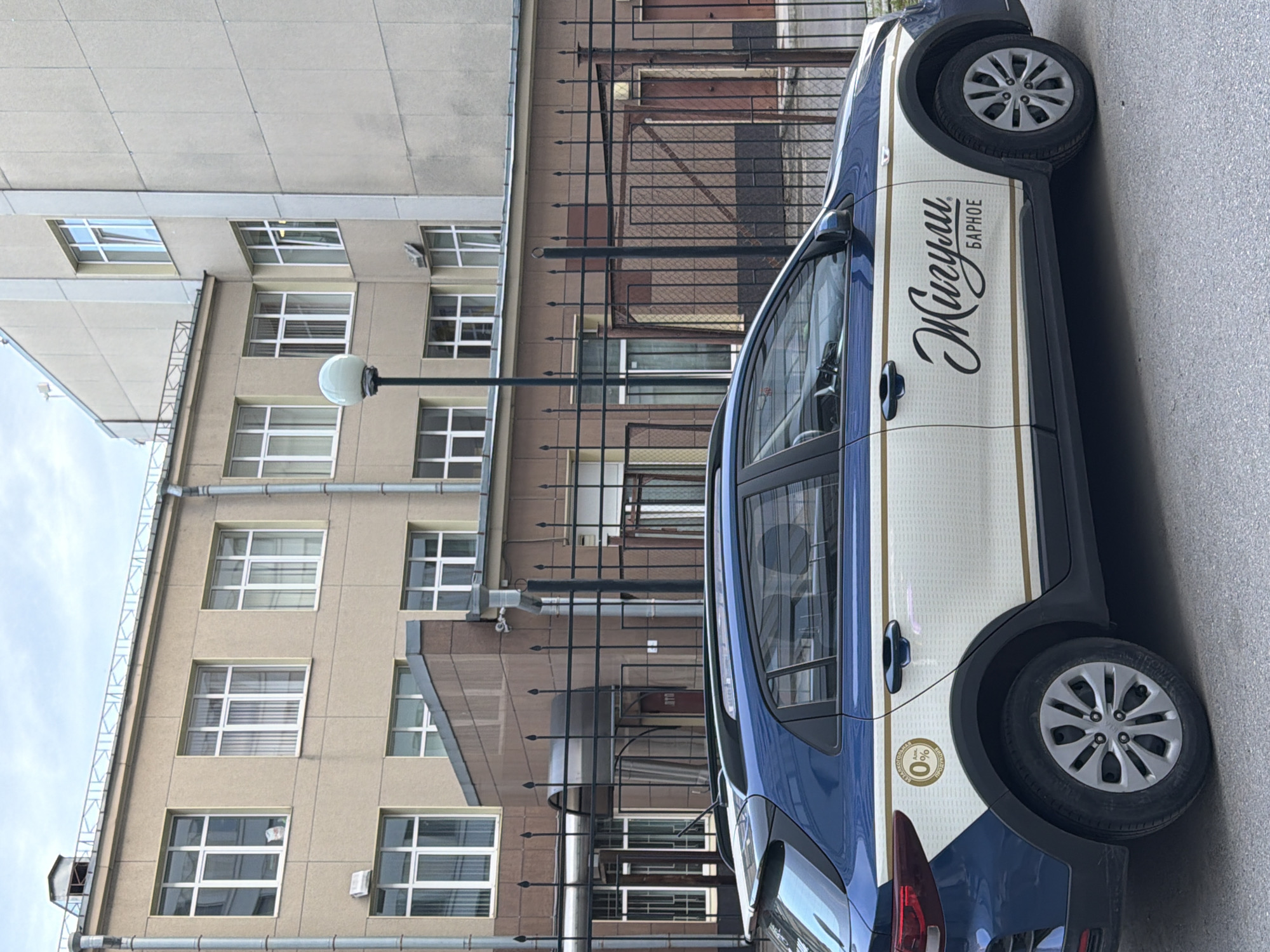 A blue and white Kia Rio X-Line, branded with "Жигули БАРНОЕ" (Zhiguli Barnoe) and "БЕЗАЛКОГОЛЬНАЯ 0% АЛК. ПРОДУКЦИЯ" (Non-alcoholic 0% Alc. Product), is parked on an asphalt street in Saint Petersburg, Russia. The vehicle features a dark blue upper body, a white lower body with horizontal gold pinstripes, and black script lettering on the side. Its wheels are silver multi-spoke alloys.
In the background, a multi-story building complex is visible behind a black metal fence with pointed finials. The building on the left is light brown with numerous rectangular windows featuring white frames, some of which are partially open. A metal downspout runs vertically along its left edge, and a decorative metal railing is present on its roofline. Below this facade, a darker brown section with a rounded awning covers an entrance marked with a red sign displaying "37Б." To the right, a taller, lighter grey building with vertical white structural elements and fewer windows extends upwards. A spherical white lamp fixture on a black lamppost stands between the fence and the buildings. The sky is overcast and grey. No individuals are present in the frame.