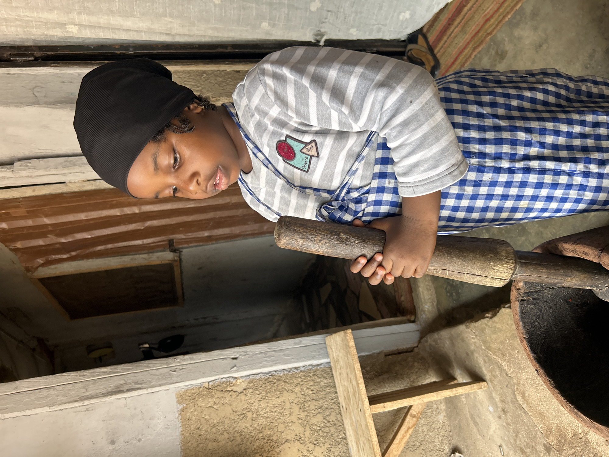 In Jos, Nigeria, a young girl wearing a black head covering, a striped shirt, and a blue and white checkered apron is using a large wooden pestle to pound something in a dark-colored mortar. The mortar is positioned on the ground. A small, wooden stool is nearby. The setting appears to be indoors, possibly a kitchen or food preparation area. The walls are painted in light colors, and a doorway is visible in the background.