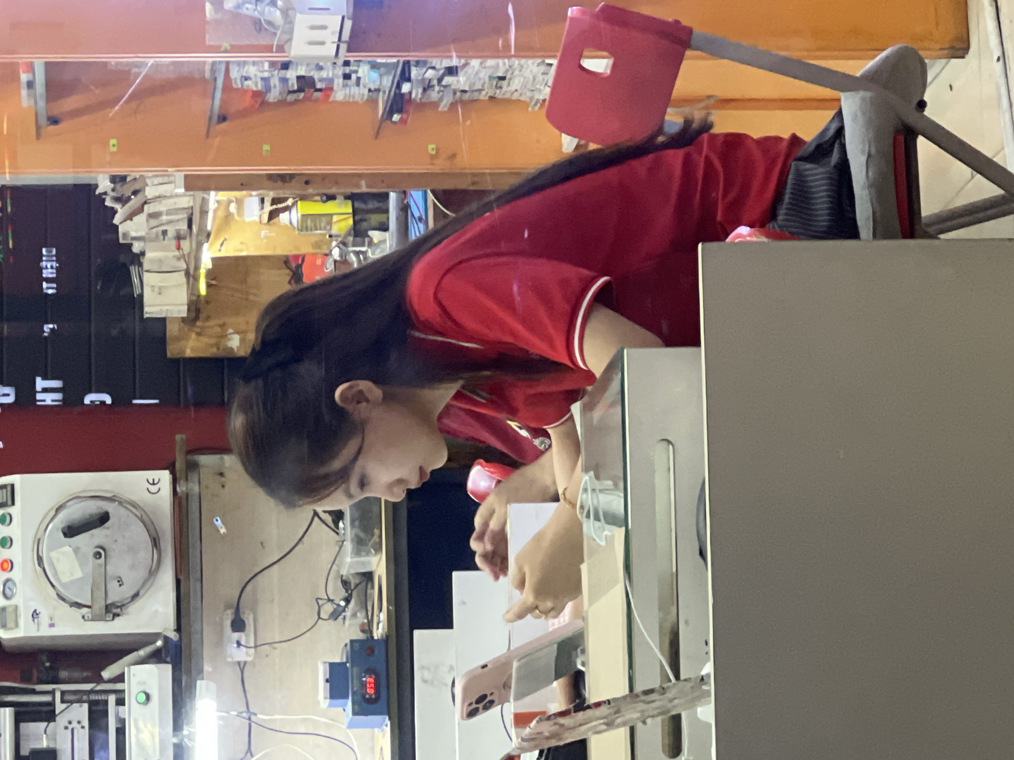 A young woman with long dark hair is seated at a desk in what appears to be an electronics repair shop. She is wearing a red short-sleeved polo shirt with white trim and a black and grey striped skirt. She is looking down at a pink cell phone held in her hands, which is resting on a white stand. The desk is cluttered with electronic equipment and tools. In the background, there is a large white machine with a circular door, labeled "CE". To the right of the woman is an orange wall with shelves displaying various items. The overall environment suggests a commercial space focused on electronic device services.