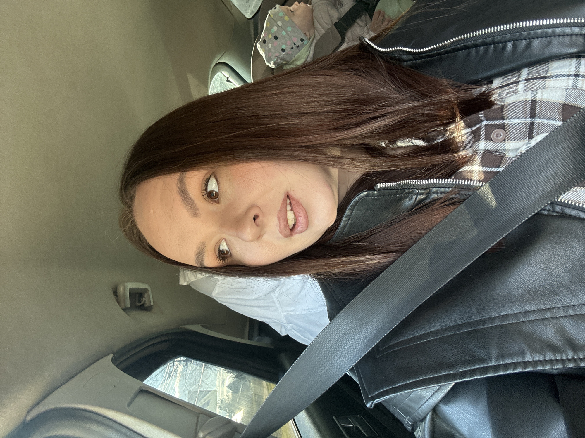 A selfie captures a young woman seated in the front passenger seat of a car, with a child visible in the back. The woman, with long dark brown hair, fair skin, and brown eyes, looks directly at the camera with a wide-eyed, slightly surprised or playful expression and an open-mouthed smile showing her teeth. She wears a black faux leather jacket with prominent silver zippers over a light brown and white plaid shirt, and a white undershirt. A black seatbelt stretches diagonally across her chest and lap.
In the upper right background, a baby or small child is partially visible in a car seat, covered by a light-colored blanket or wrap adorned with multicolored polka dots. The car's interior appears to have a light grey or tan headliner. Through the window on the right, a blurry outdoor scene suggests bare tree branches, indicating an autumn or winter setting during daylight hours. The overall ambiance is casual. No visible text is present in the image. The setting is a car interior, with the provided context placing it in Chaikovsky, Russia.