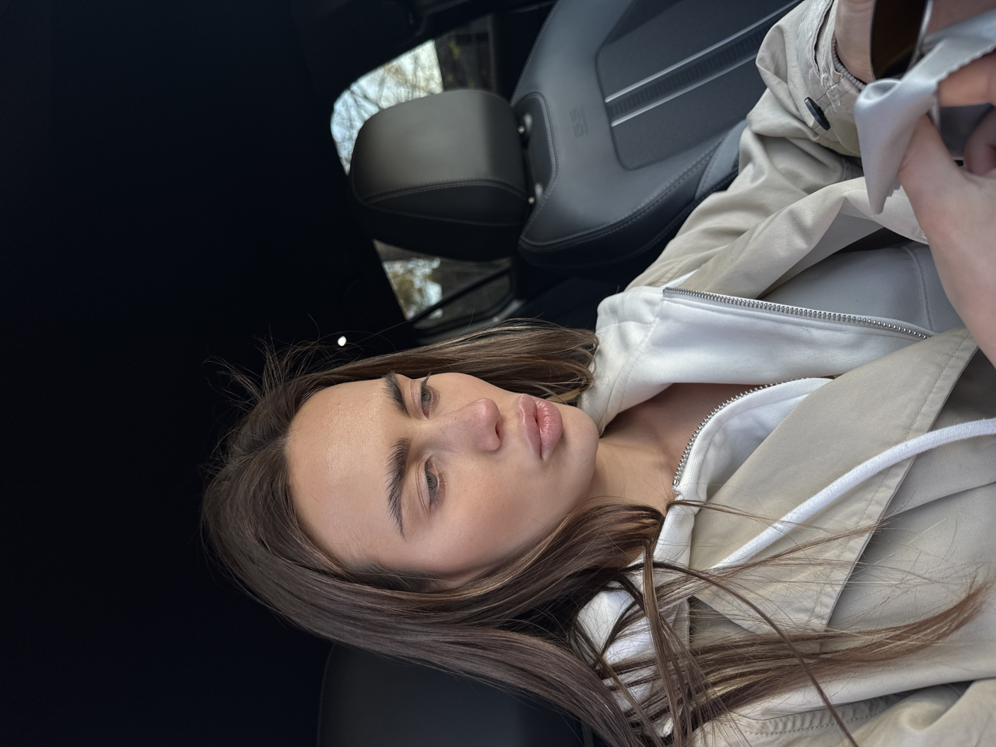 A young woman with long brown hair is seated in a black car, wearing a white top and a beige jacket. The car's interior is visible, including a black leather seat with white stripes. The location is not specified in the image or crops, nor is the city of Białobrzegi, Poland.