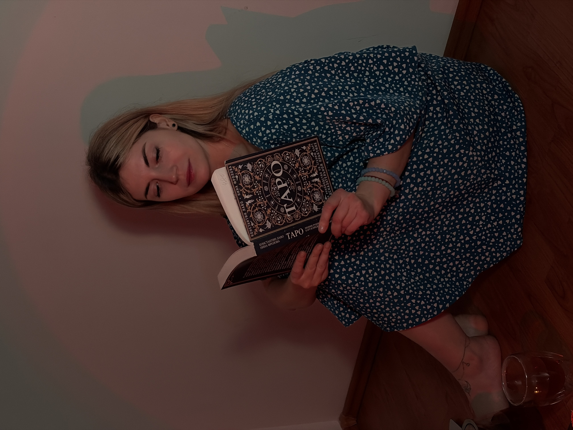 A woman is reclined against a pale-colored wall in a corner, wearing a blue dress with white heart-shaped patterns. She is holding a book titled "TAPO," written in a Cyrillic script, and appears to be asleep or resting with her eyes closed. The floor is made of wooden planks. A glass with a dark liquid sits on the floor near her bare feet, one of which has a tattoo on the ankle. She wears bracelets on her wrist. The lighting is dim and warm, suggesting an indoor setting, not specific to Budva, Montenegro.