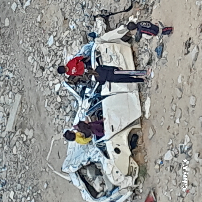 The image depicts a desolate, rubble-strewn landscape in Khan Yunis, Palestinian Territory. A severely damaged white four-door sedan occupies the center of the frame, resting on uneven ground covered in fragmented concrete and debris. The car's roof is heavily crushed, and its body panels are mangled and dislodged, with missing wheels. Five children are present, interacting with the wreckage.
Two children are on the car's mangled roof: one in a yellow long-sleeved top and dark bottoms near the left side, and another in a red long-sleeved top with a white graphic and dark bottoms towards the right. A third child, wearing a dark purple garment, is partially visible within the car's interior, possibly leaning through a window frame. A fourth child, dressed in a dark tracksuit with white stripes along the legs, stands upright next to the car's passenger side, facing towards the vehicle. A fifth child, wearing a dark top with red and white detailing on the sleeves and dark bottoms, stands further to the lower right, facing the car. The children appear to be exploring or playing on and around the wrecked vehicle. The bottom left corner of the image contains a "Galaxy A14" watermark.