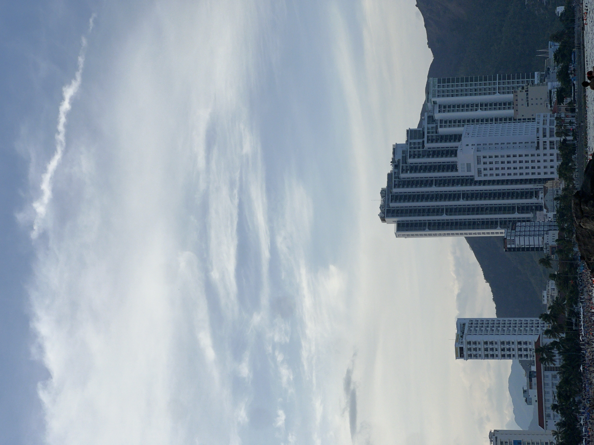 The image depicts a vibrant coastal scene, rotated 90 degrees clockwise from its natural orientation. When viewed upright, a wide expanse of light blue sky with scattered, wispy white clouds dominates the upper and left portions.
In the mid-ground and right, a dense cluster of modern, white high-rise buildings lines a shoreline, extending towards a dark, imposing mountain range that forms the backdrop. These multi-story structures feature numerous balconies, suggesting hotels or residential complexes. One prominent building displays the vertical text "SCANIA" on its facade, and another shows "YDCCO".
Along the immediate right edge, a body of water, likely the ocean or a bay, is visible. The shoreline beside the water is teeming with a crowd of people, appearing as numerous small figures, indicative of a busy beach or resort area. A few larger figures are distinguishable closer to the foreground near the water's edge, and some green palm trees dot the landscape among the buildings.
The activity taking place suggests a leisurely day at a popular coastal destination, with people gathered by the water. The scene unfolds during daytime, under bright conditions, with clear skies and good visibility. The presence of tall buildings, a mountain, and a crowded shoreline strongly points to a bustling resort city like Bac Nha Trang Ward, Vietnam, as indicated by the context.