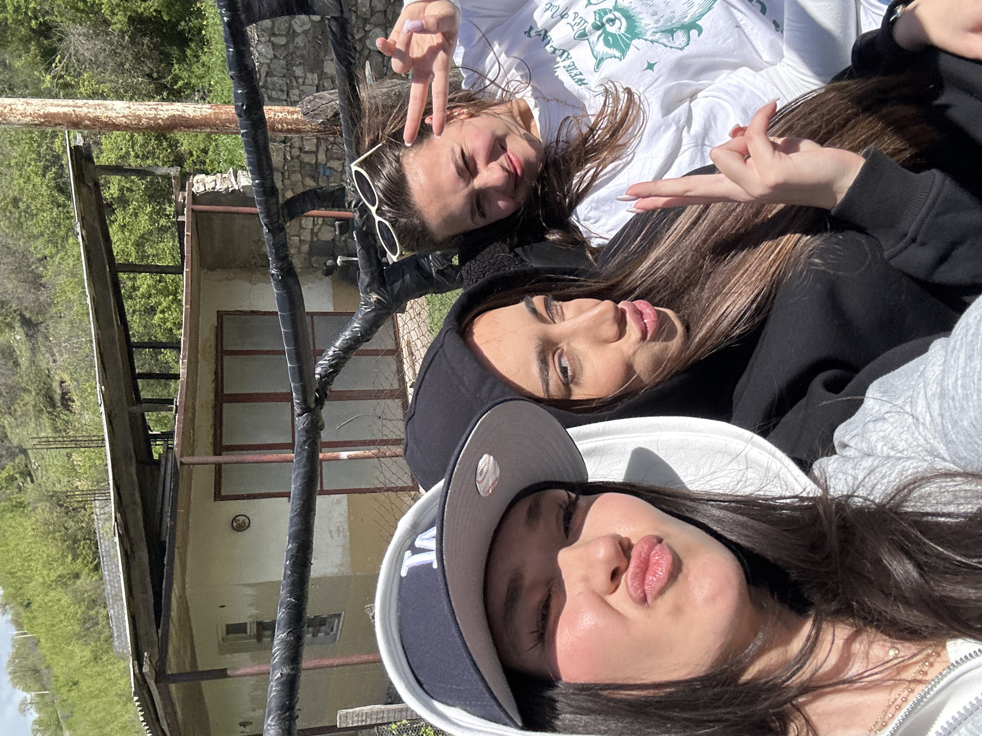 Three young women are taking a selfie, with part of a building and scaffolding visible in the background. The woman on the top is wearing a white long-sleeve shirt with a graphic design, and sunglasses. She is making a peace sign with her left hand. The woman in the middle is wearing a black hooded jacket. The woman on the bottom is wearing a white and black New York Yankees baseball cap. The building in the background appears to be old and in disrepair, with wooden window frames and a stone foundation. Green vegetation is seen beyond the building. The scaffolding is made of metal pipes wrapped in black material. The photo was taken during the day in a location with abundant natural light, possibly Haghartsin, Armenia.
