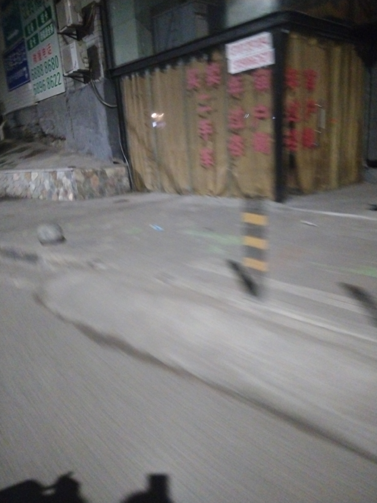The image shows a street scene in Jiulongpo District, China at night. A building with a sign featuring Chinese characters and the numbers 6888 8680 and 6896 8622 is visible on the left. Next to it is a closed storefront or building section, with curtains or drapes covering the windows. The storefront has writing or decorations on the glass. The foreground consists of a paved street, and the image is blurry, indicating motion. A traffic bollard is to the center right. The overall lighting is dim, contributing to the nighttime atmosphere.