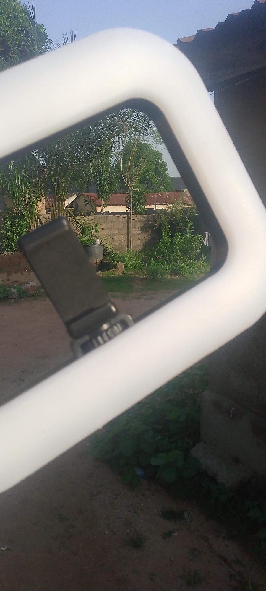 The image depicts a close-up view of a white, tubular structure, possibly part of a frame or railing, which dominates the foreground. Through an opening in this structure, a background scene is visible. This background includes a wall constructed of grey concrete blocks, overgrown with green vegetation. Beyond the wall, there are buildings with corrugated iron roofs, suggesting a residential or semi-rural area. Trees and other greenery are also present. In the mid-ground, a black object, which appears to be a part of a device or accessory, is attached to the white structure. This object has a textured surface and metallic elements, resembling a clasp or a buckle. The sky is visible above, appearing light blue. The overall setting suggests an outdoor environment, possibly in Jalingo, Nigeria, given the architectural style and vegetation. The lighting indicates it is daytime, with the sun casting shadows.