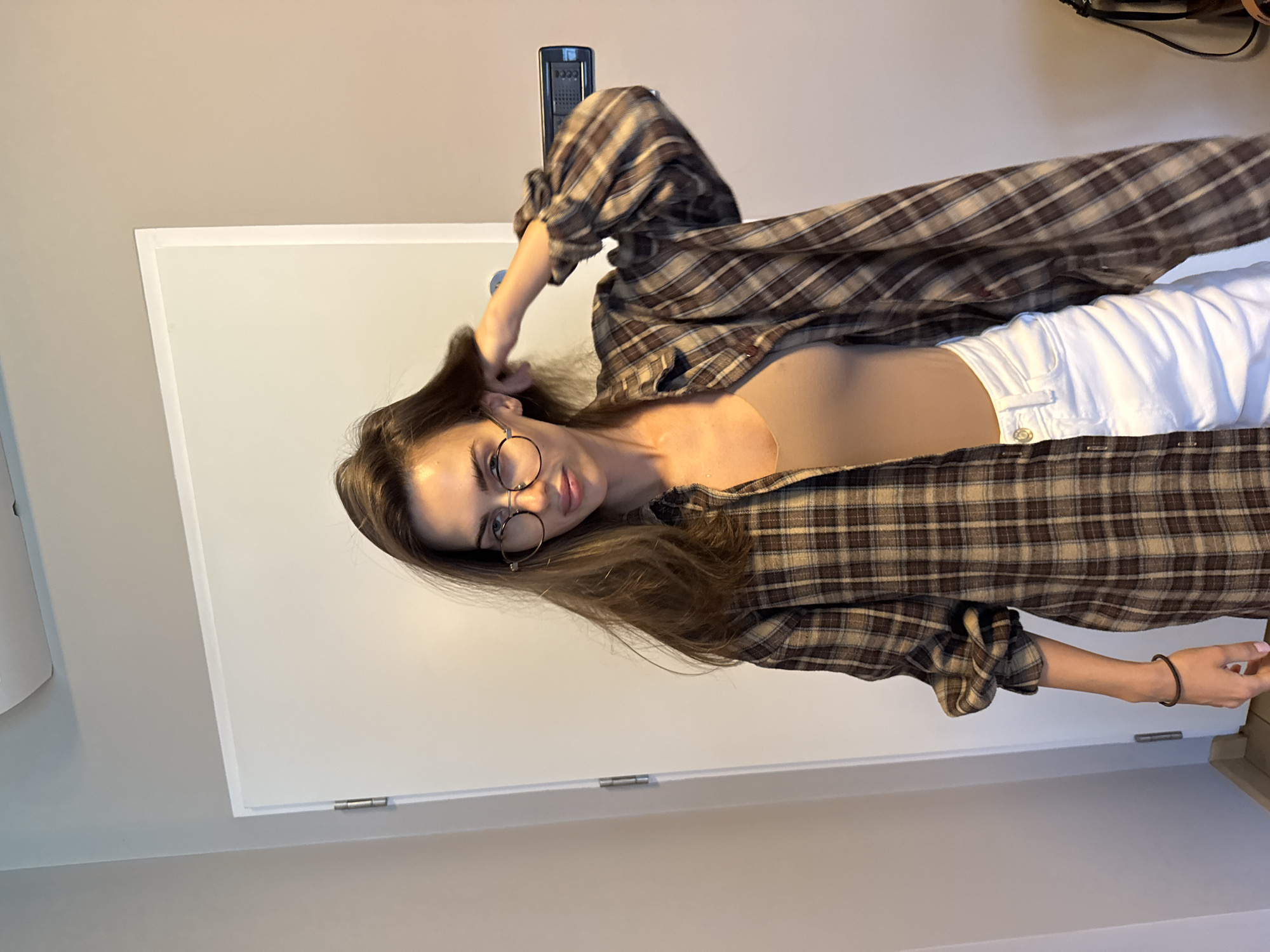 A young woman stands in a room, clad in a brown plaid shirt over a light top and white pants. She wears circular-framed glasses and touches her brown hair with her left hand. A black bracelet adorns her right wrist. The setting includes a white door in the background and a gray wall. An electronic device hangs on the wall above her. A glimpse of furniture is visible to the right. The location appears to be an indoor setting. The city is unknown.