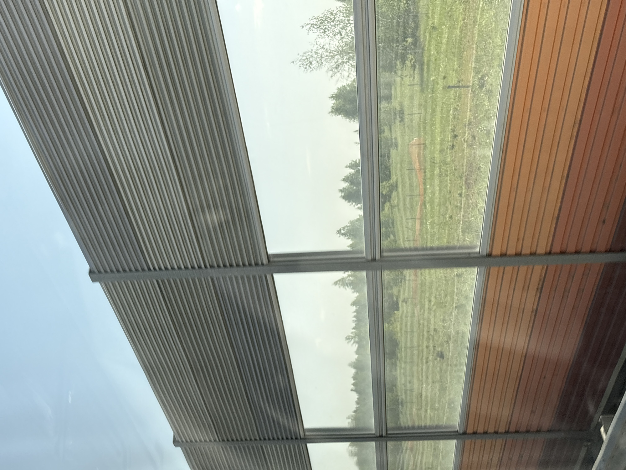 The image shows a partial view of a structure, possibly a bus stop or a covered walkway, featuring a corrugated metal roof on the left and a wooden slat roof on the right. Between the two roof sections are glass windows offering a view of a green, grassy field with trees. The scene is slightly overexposed, obscuring detail in the landscape. No people or other subjects are visible. It is impossible to confirm the location of the city of Сапуголи, Russia from this image.