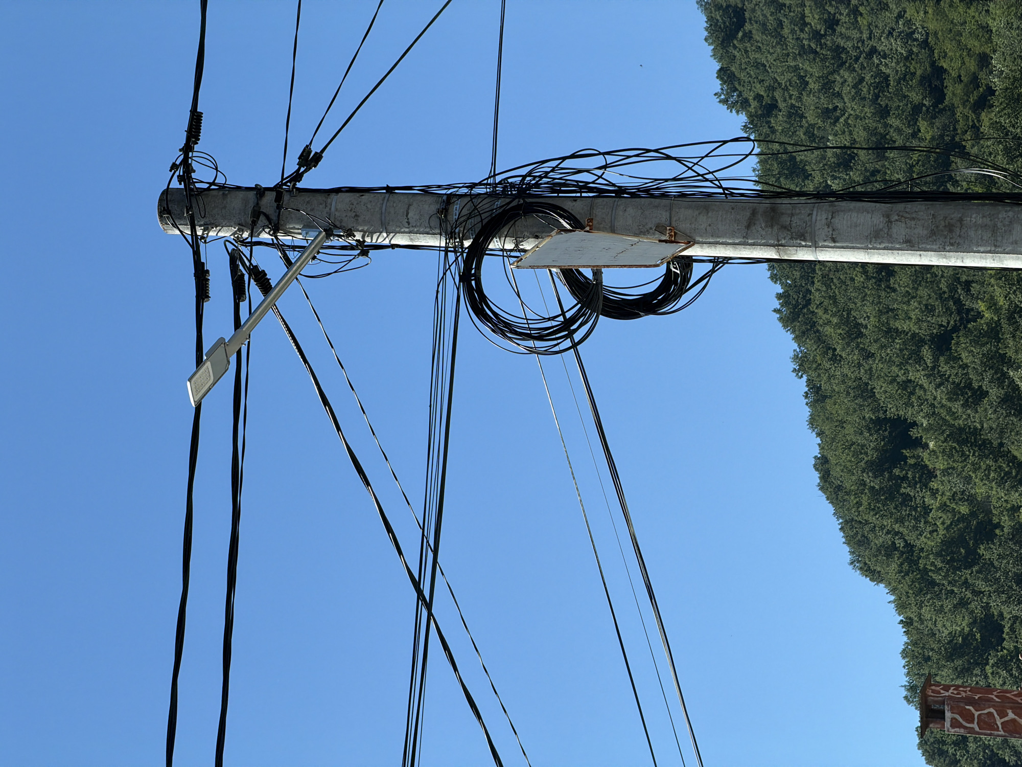 The image shows a utility pole in Beli Osam, Bulgaria, against a clear blue sky. The concrete pole is heavily laden with numerous black electrical and communication wires, some of which are coiled around the pole. A street lamp is attached to the upper section of the pole. In the background, a lush green, tree-covered hillside is visible, suggesting a natural or semi-rural setting. A portion of a decorative chimney, with a textured surface, is present in the lower left corner of the frame. There are no people or animals visible in the image.