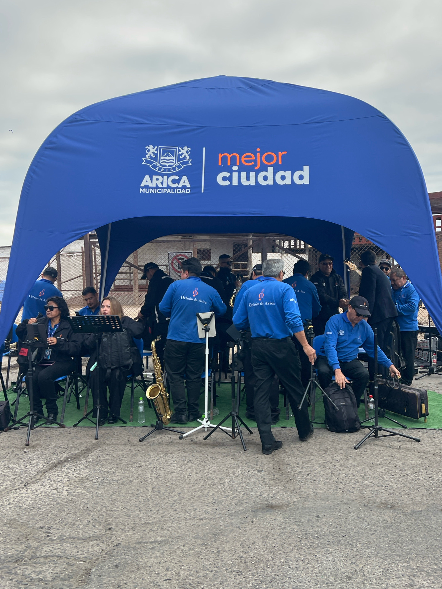 A group of musicians, dressed in blue shirts with the "Orfeón de Arica" logo, are gathered under a large blue tent with "ARICA MUNICIPALIDAD" and "mejor ciudad" branding. The scene appears to be outdoors, possibly a public square or street in Arica, Chile, given the prominent municipal branding. The musicians are preparing for a performance, with instruments such as a saxophone visible. Some are seated on chairs, while others are standing. The ground is paved and appears somewhat worn. The sky is overcast, suggesting a cloudy day. The overall atmosphere suggests an organized event or performance sponsored by the municipality of Arica.