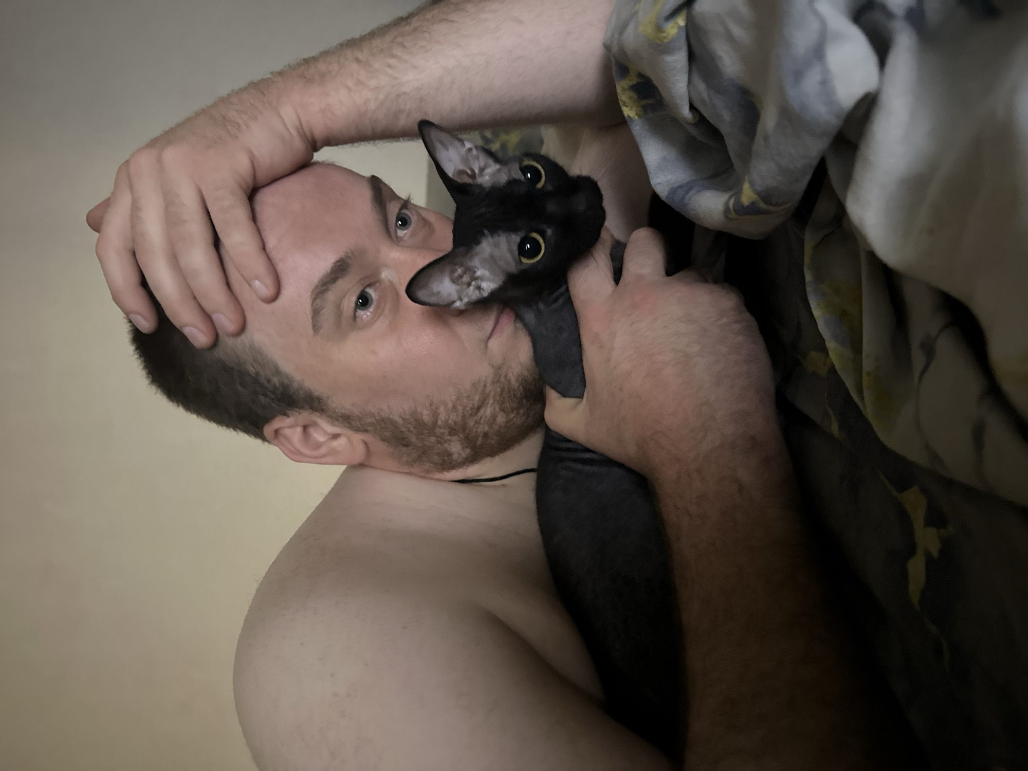 A close-up indoor shot features a shirtless man holding a dark gray or black cat against his face. The man has a beard, short hair, and light skin. His left hand is on his forehead. The cat has large yellow eyes and is facing the camera. The background includes bedding or a blanket with a pattern in shades of gray and yellow, and a light-colored wall. The location is Chaikovsky, Russia.