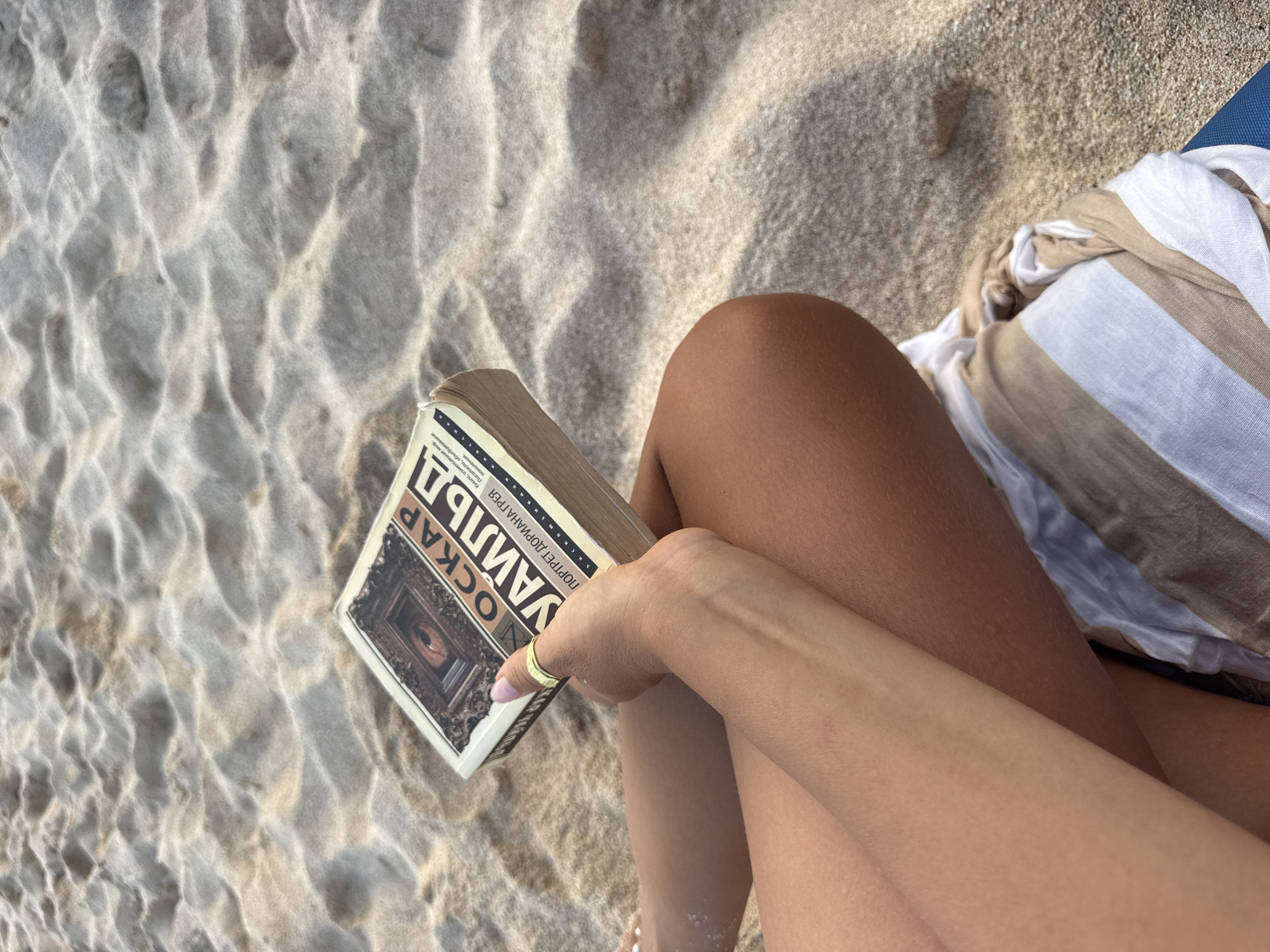 A close-up, overhead shot shows a person sitting on a sandy beach in Kuta Selatan, Indonesia. The person's legs are crossed, and they are holding an open book titled "Оскар Уайльд" with a portrait of Dorian Gray on the cover. The book is held in the person's right hand, which is adorned with a gold ring on the index finger. The sand on the beach has rippled patterns created by wind or water. A striped beach towel is visible under the person's legs. The lighting suggests it is daytime, with sunlight casting shadows on the sand.